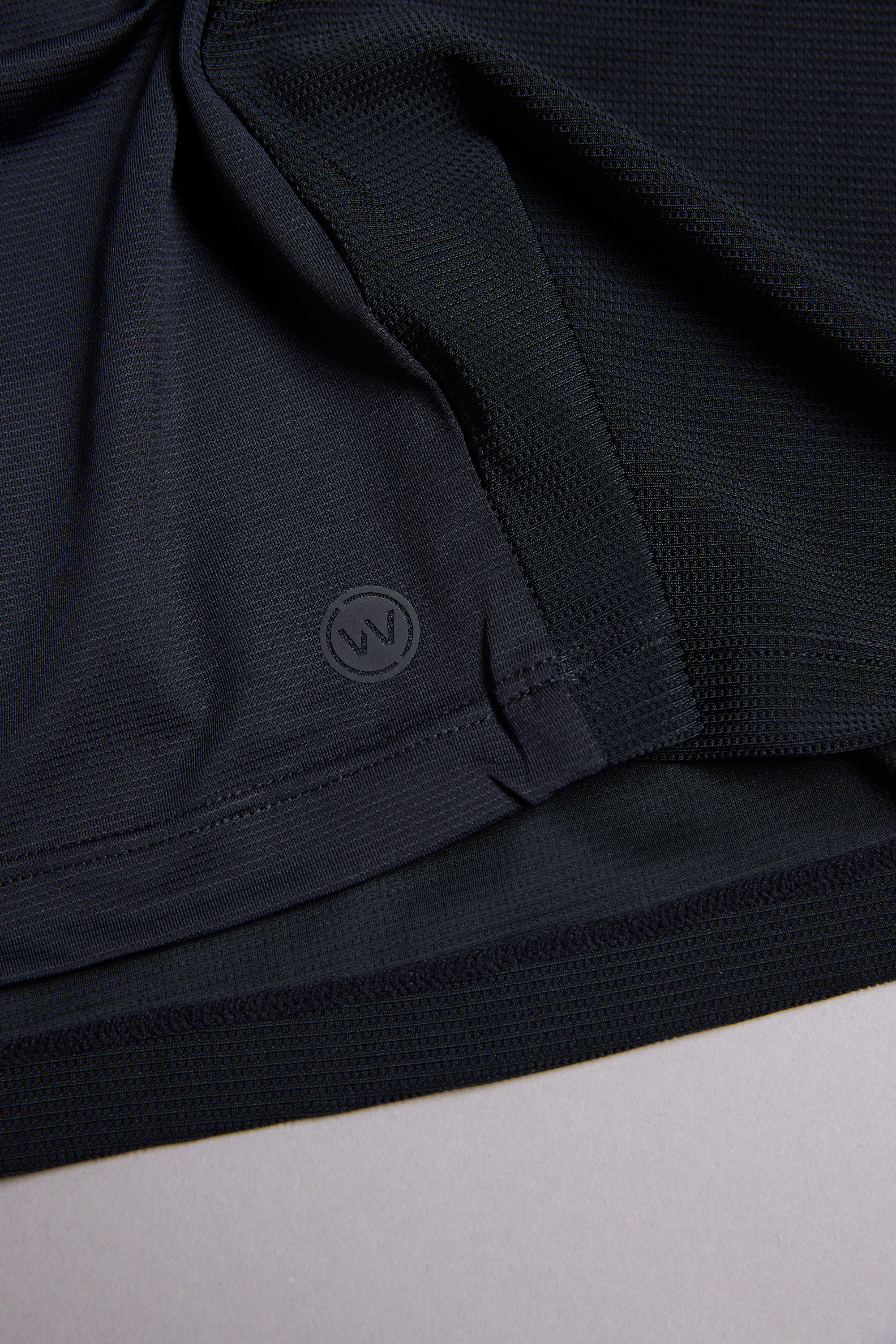 Close-up of the WearEver Pulse Tee's dark, textured performance fabric with a stitched hem and a small circular WearEver "W" logo near the edge, lying flat on a light surface.