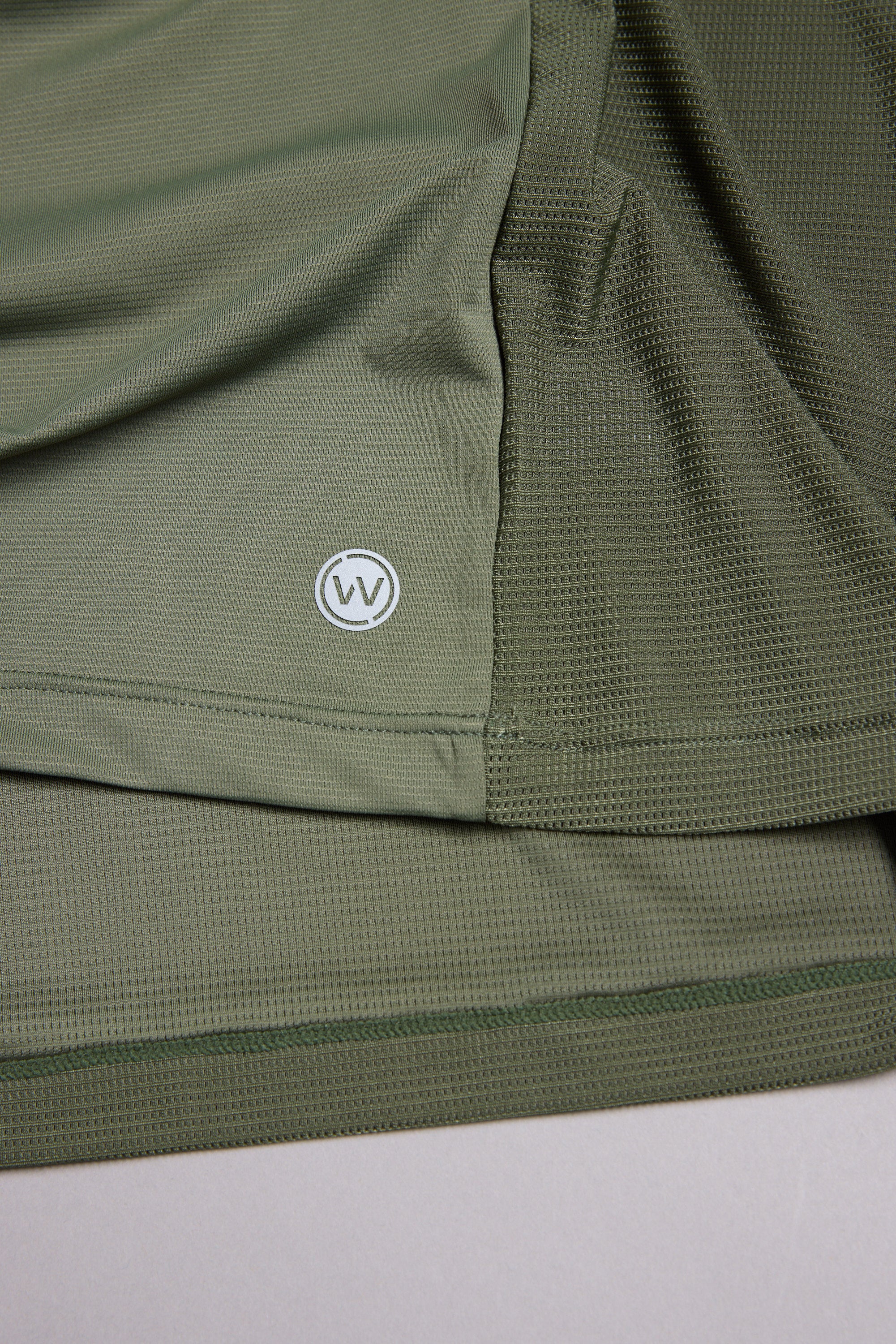 A close-up of olive green mesh fabric on the WearEver Pulse Tee by WearEver, a moisture-wicking performance t-shirt featuring a small circular overlapping V logo near the hem.