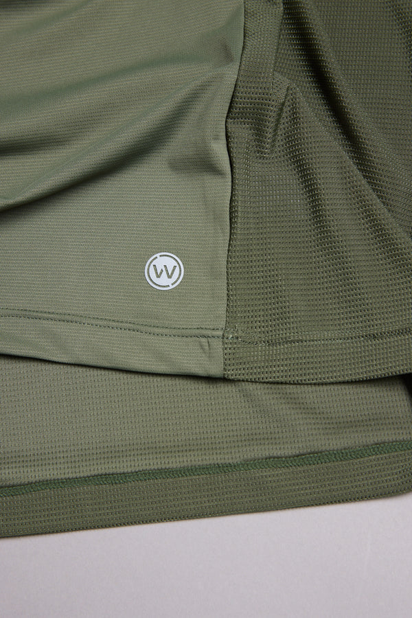 A close-up of olive green mesh fabric on the WearEver Pulse Tee by WearEver, a moisture-wicking performance t-shirt featuring a small circular overlapping V logo near the hem.
