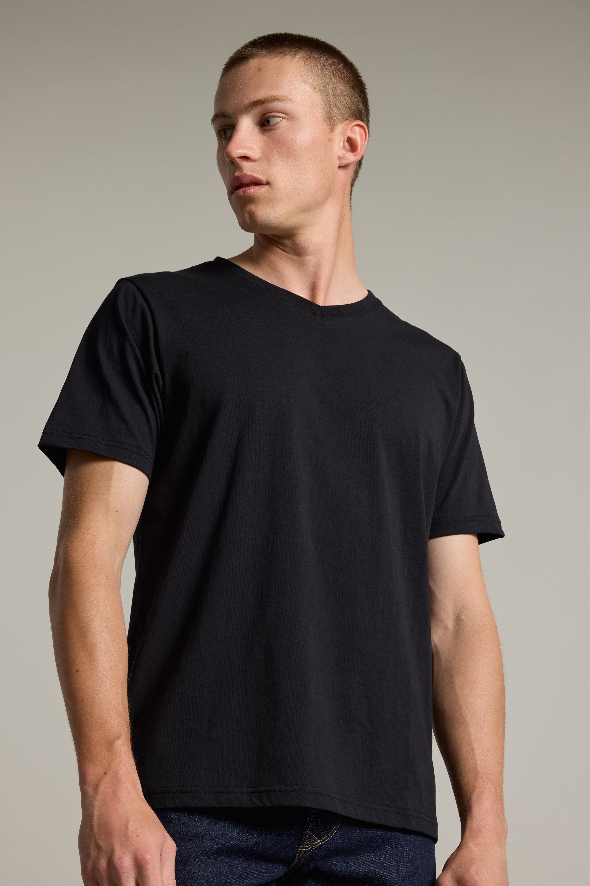 A young man with short hair wears the Barkers Organic Cotton V Neck Tee and dark jeans, standing against a neutral background and looking left with a serious expression—a timeless essential for any wardrobe.
