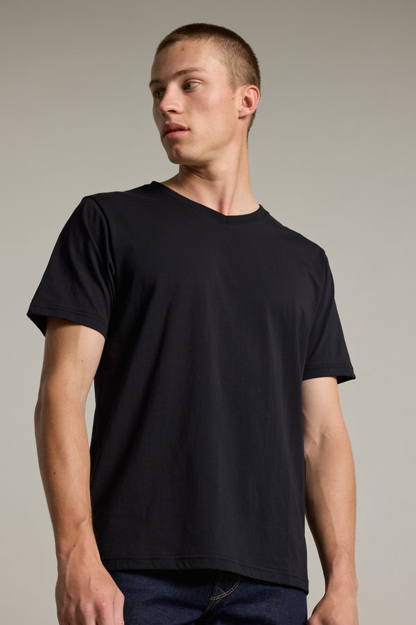 A young man with short hair wears the Barkers Organic Cotton V Neck Tee and dark jeans, standing against a neutral background and looking left with a serious expression—a timeless essential for any wardrobe.