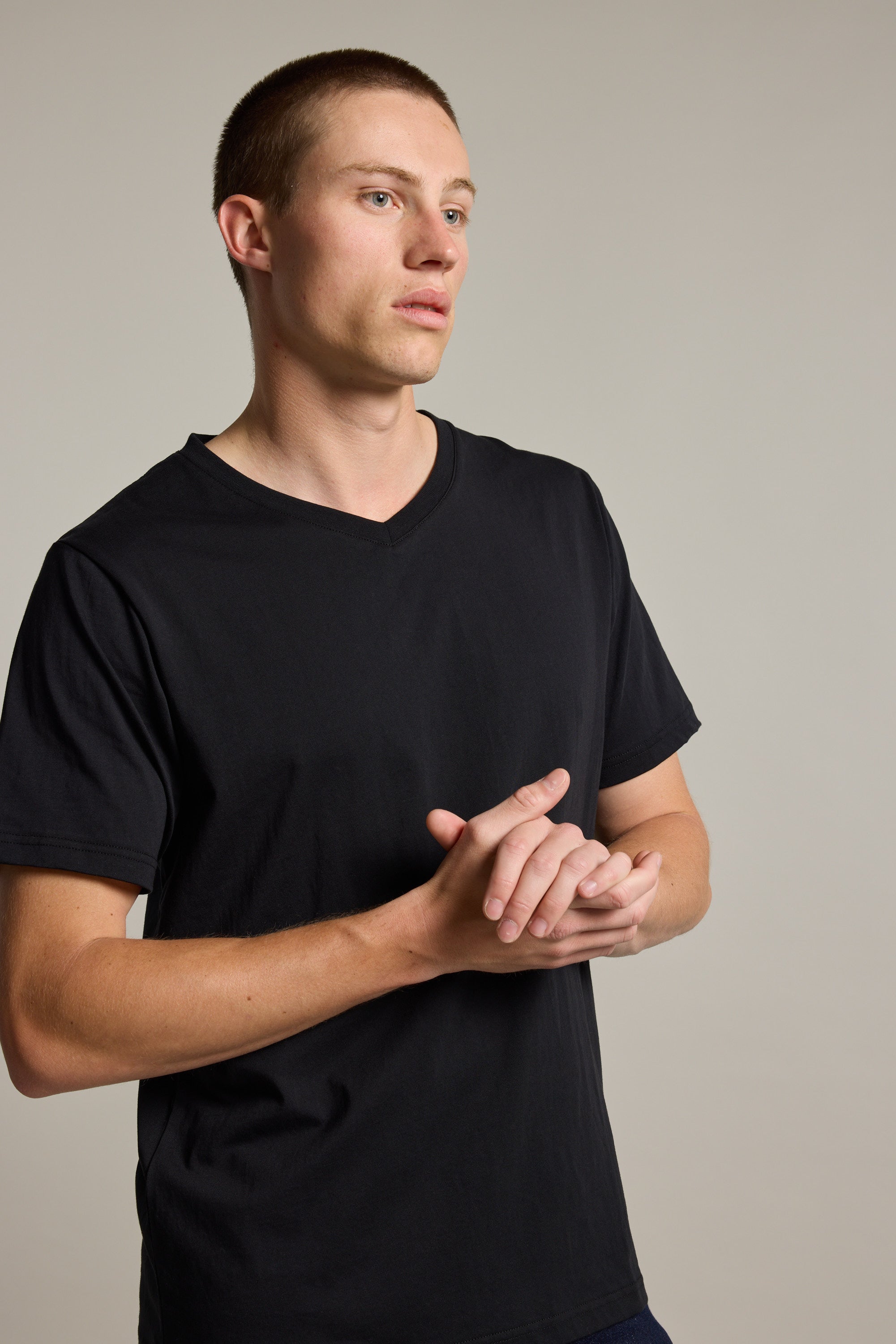 A young man with short hair stands against a plain light background, wearing the Barkers Organic Cotton V Neck Tee—an everyday essential—as he looks to the side with his hands clasped.