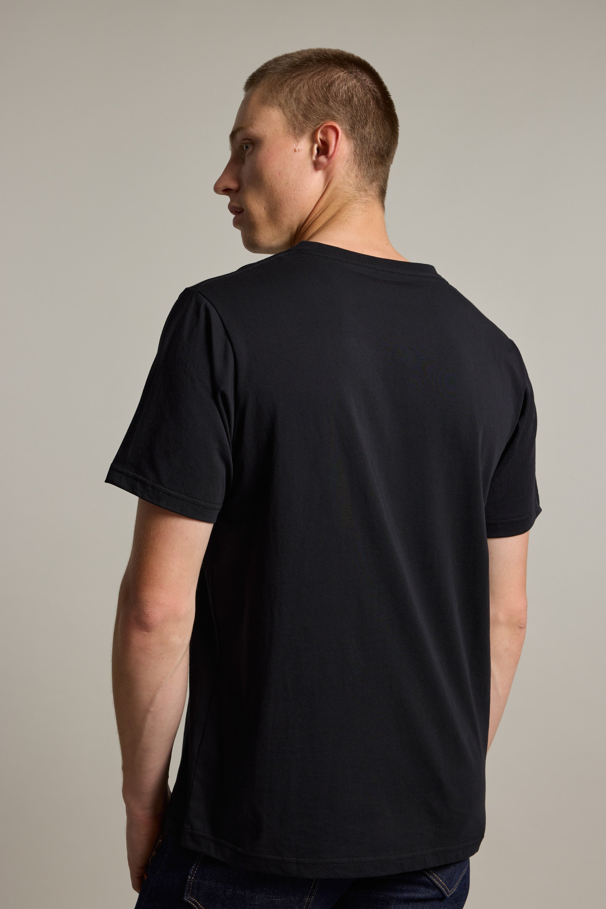A man with short brown hair, wearing the Barkers Organic Cotton V Neck Tee in black and dark jeans, stands facing away from the camera and looks over his left shoulder, set against a plain, light background.