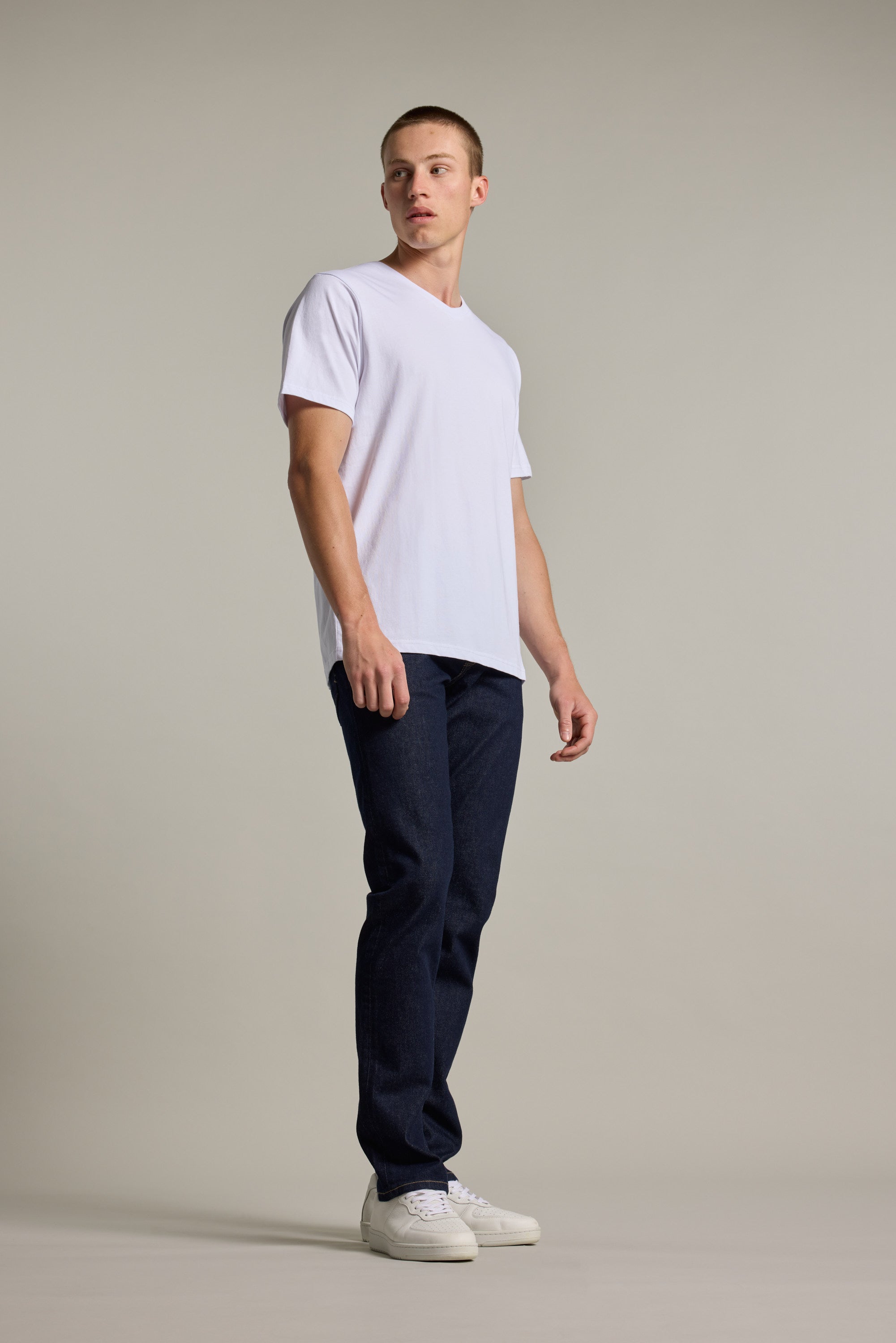 A young man stands against a plain background in the Barkers Organic Cotton V Neck Tee, paired with dark blue jeans and white sneakers. He gazes slightly to the side with a neutral expression.