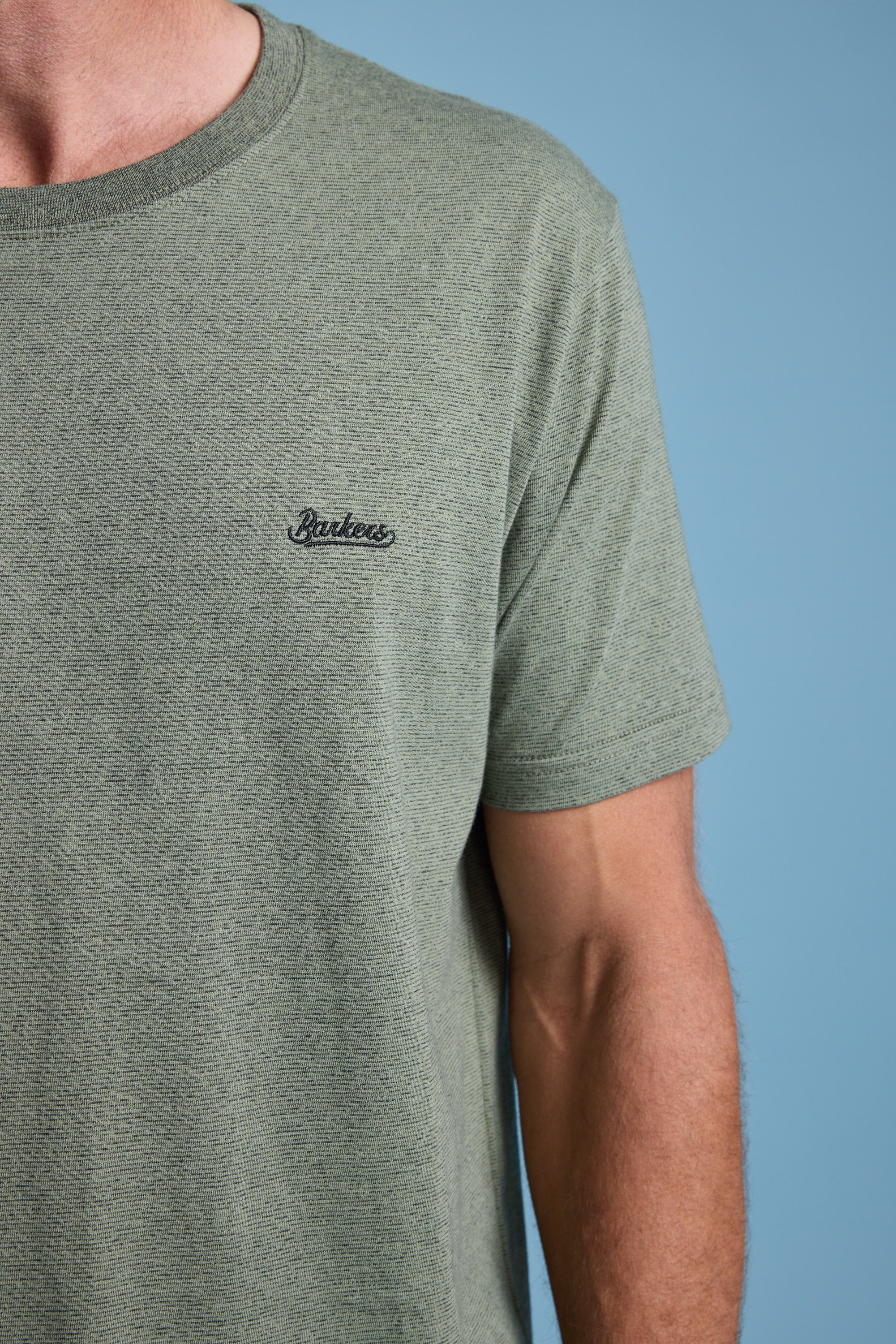 A person in a Barkers Bayside Emb Tee—light olive green, organic cotton, short-sleeved with a small chest logo—stands against a solid light blue background. Only the upper torso and part of one arm are visible.