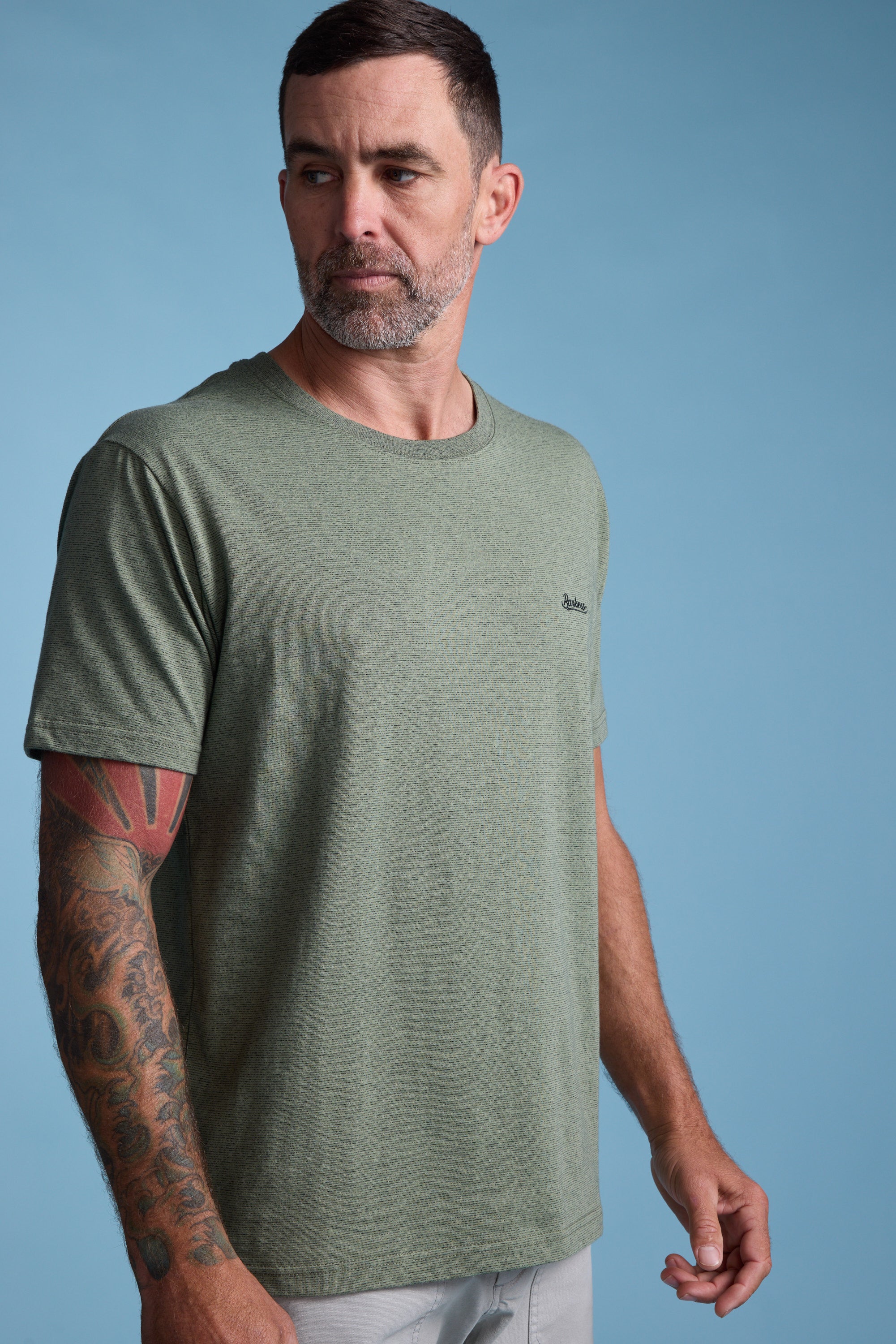 A man with short dark hair and a beard, wearing the Barkers Bayside Emb Tee in light green and light-colored pants, stands against a blue background. He has a tattoo sleeve on his left arm and looks slightly to the side.