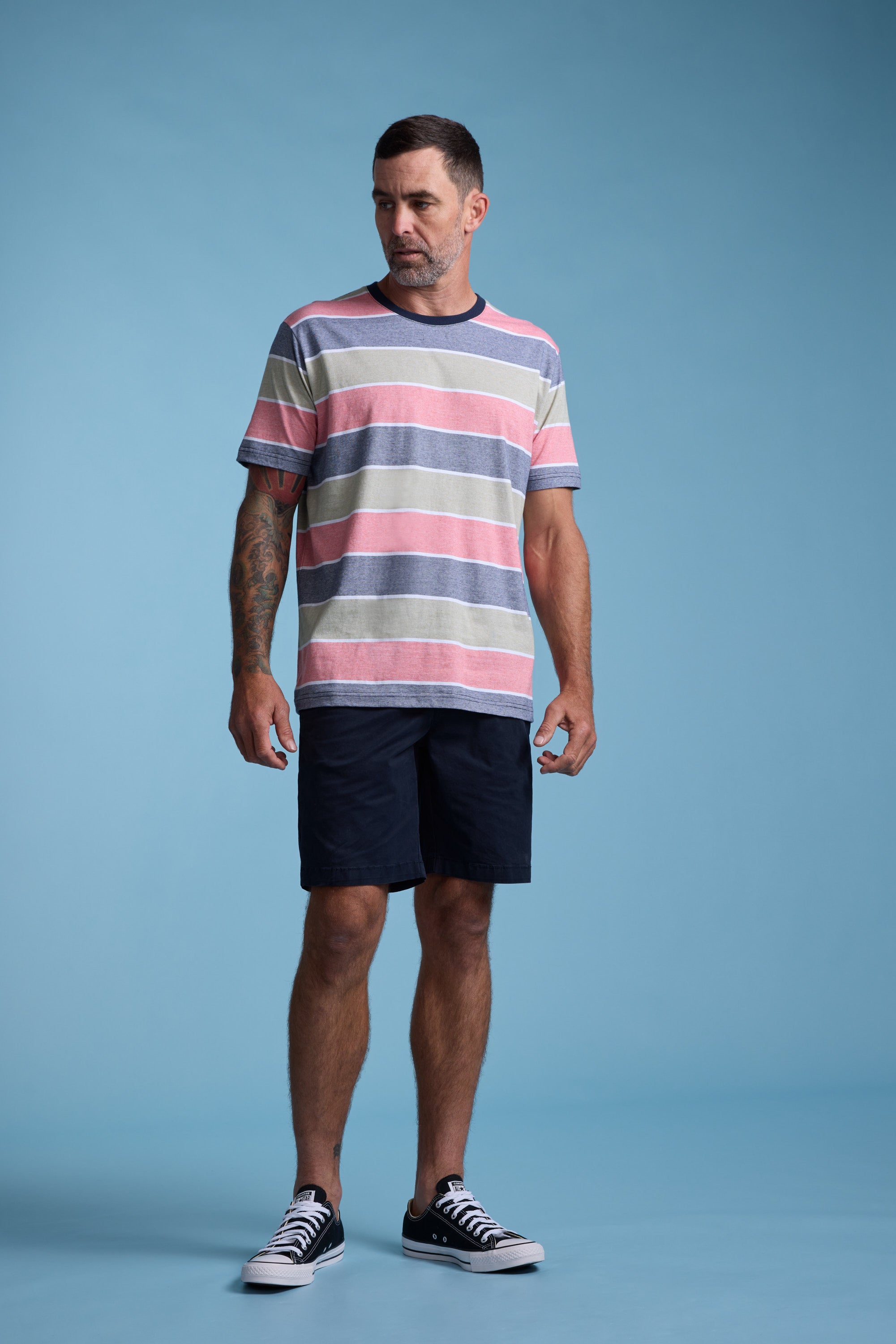 A man with short hair, a beard, and tattoos on his right arm stands against a light blue background wearing the Barkers Asbrooke Stripe Tee, paired with dark shorts and black sneakers with white soles.
