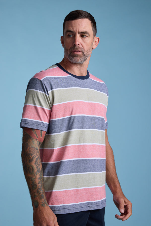 A man with short dark hair and a beard wears the Barkers Asbrooke Stripe Tee—a pink, gray, and white striped shirt—against a plain light blue background. He has a tattooed arm and glances slightly to the side.