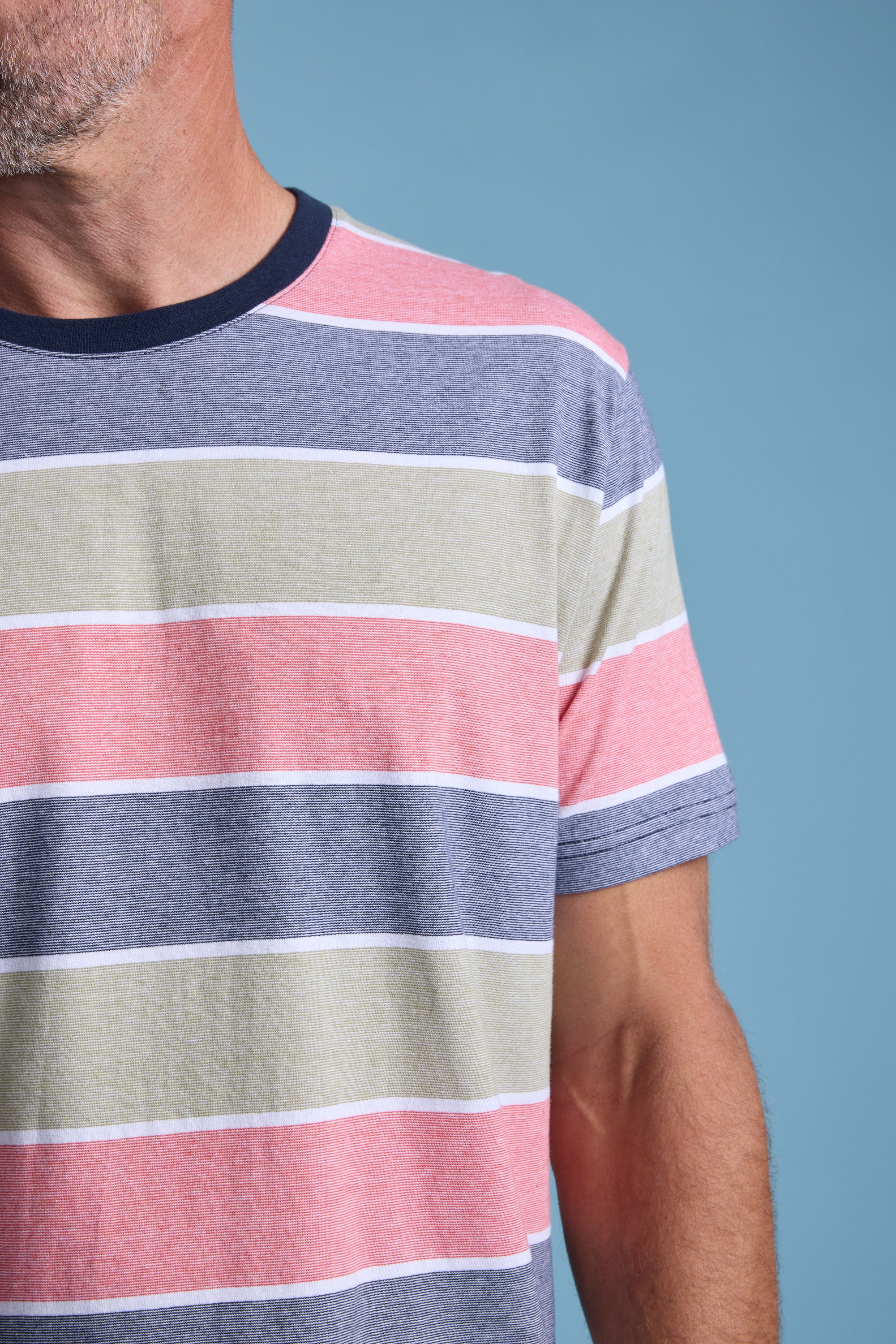 A close-up of a person wearing the Barkers Asbrooke Stripe Tee—a short-sleeved essential in blue, pink, green, and white stripes—against a plain blue background. Only the lower face, neck, and upper torso are visible.