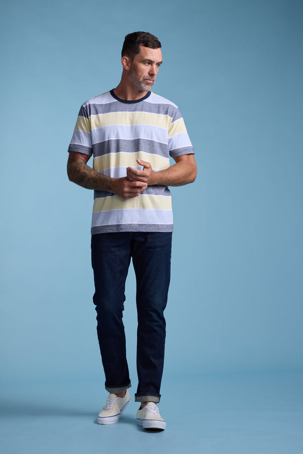 A man with short dark hair, a beard, and tattoos on one arm stands against a light blue background wearing the Barkers Asbrooke Stripe Tee paired with dark jeans and light sneakers.