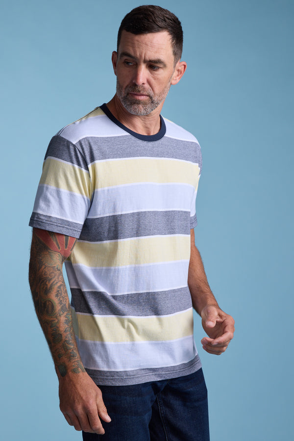 A man with short dark hair, a beard, and tattoos on his left arm wears the Barkers Asbrooke Stripe Tee—an organic cotton, regular fit shirt with yellow, gray, and white summer stripes—standing against a plain blue background and looking to his left.