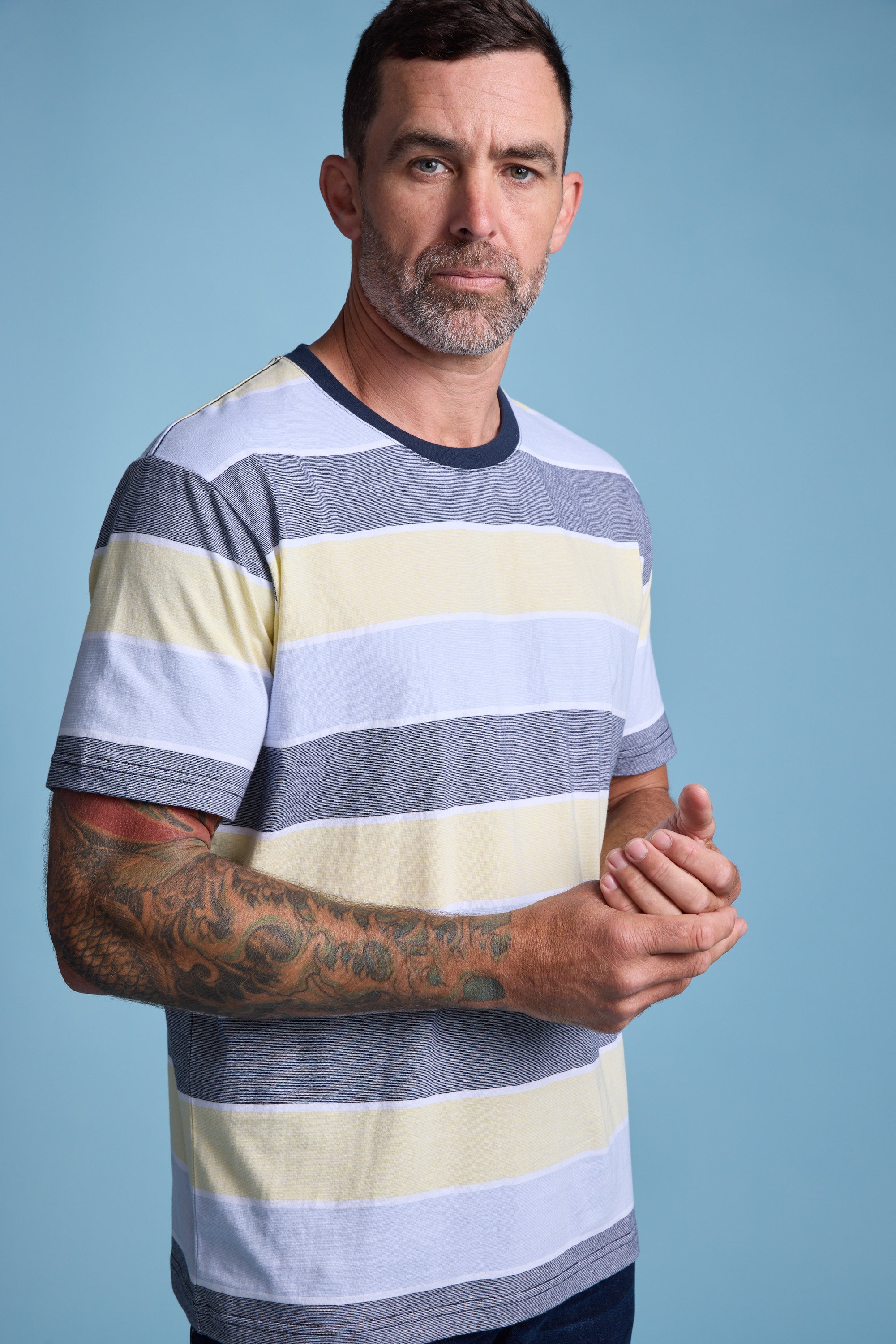 A man with short dark hair and a beard stands against a blue background, wearing the Barkers Asbrooke Stripe Tee in gray, yellow, and white. He has tattoos on his left forearm and clasps his hands together.