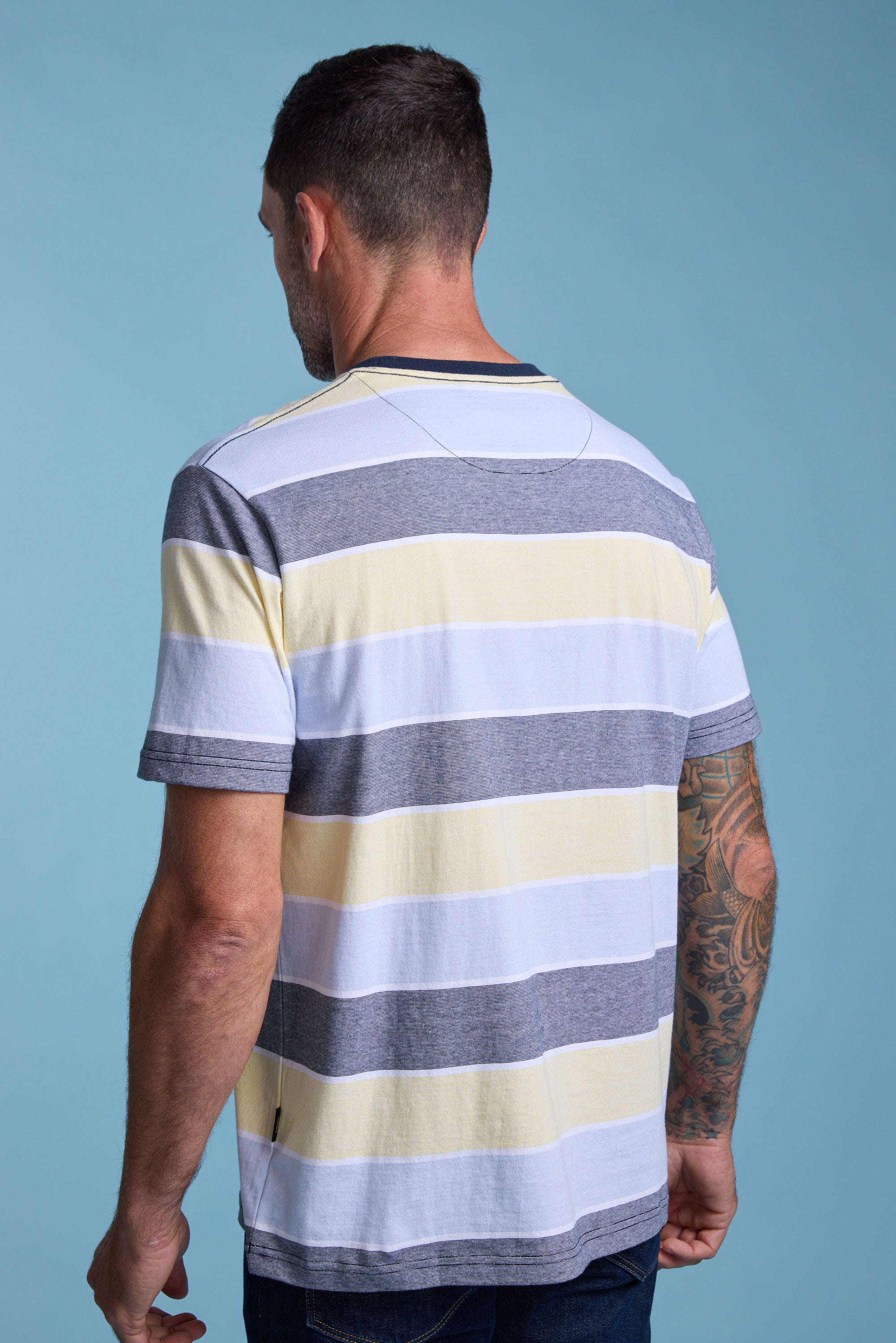 A man with short dark hair and tattoos on his right arm faces away from the camera, wearing the Barkers Asbrooke Stripe Tee—an organic cotton, regular fit shirt in gray, white, and yellow—set against a blue background.