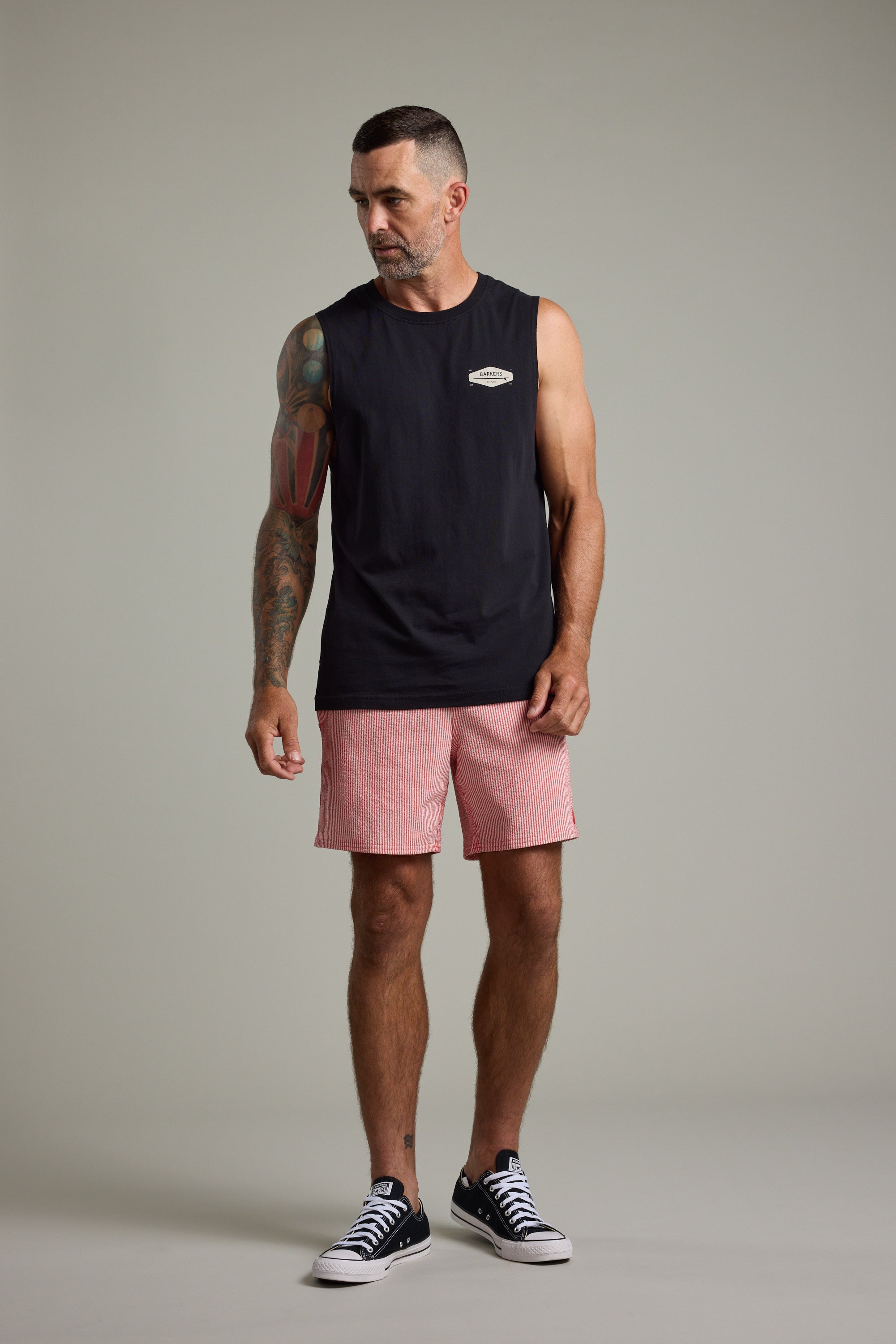A man with short hair and a beard wears the Barkers Supply Co Print Singlet by Barkers in black, paired with pink shorts and black sneakers, showing off a colorful arm tattoo while standing confidently against a plain gray background.