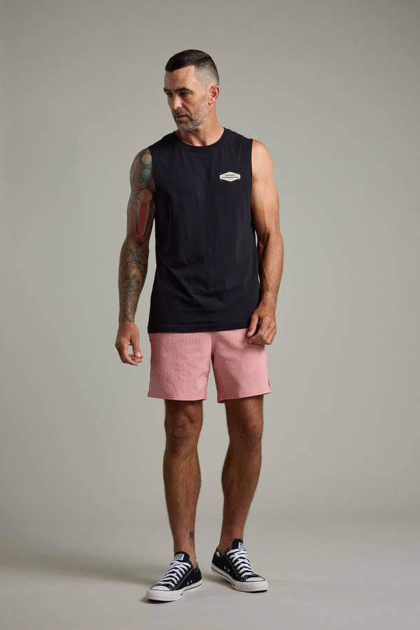 A man with short hair and a beard wears the Barkers Supply Co Print Singlet by Barkers in black, paired with pink shorts and black sneakers, showing off a colorful arm tattoo while standing confidently against a plain gray background.