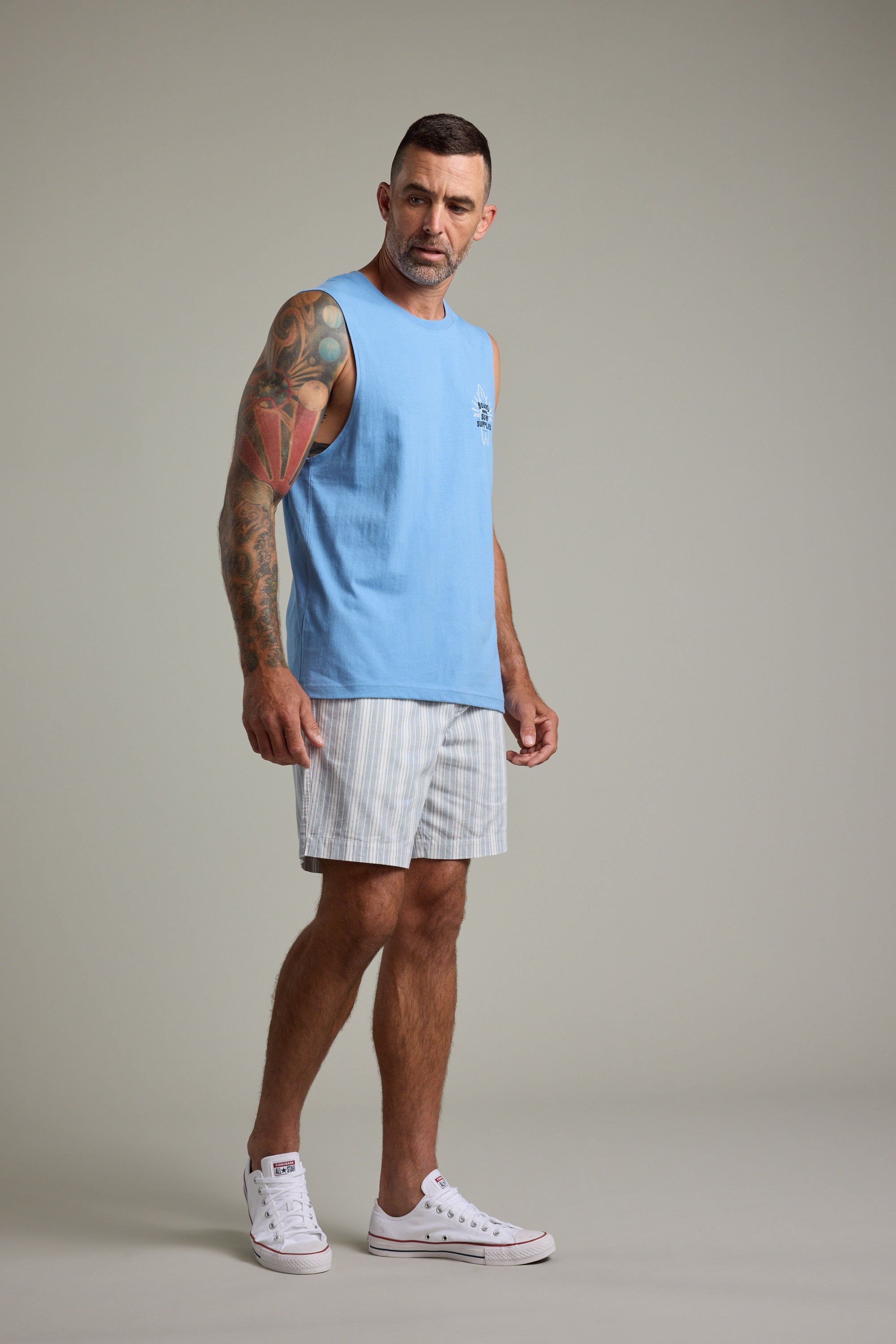 A tattooed man wears a Barkers BMC Board Riders Print Singlet in light blue, striped shorts, and white sneakers, standing on a plain backdrop and gazing slightly left—a perfect summer staple look.