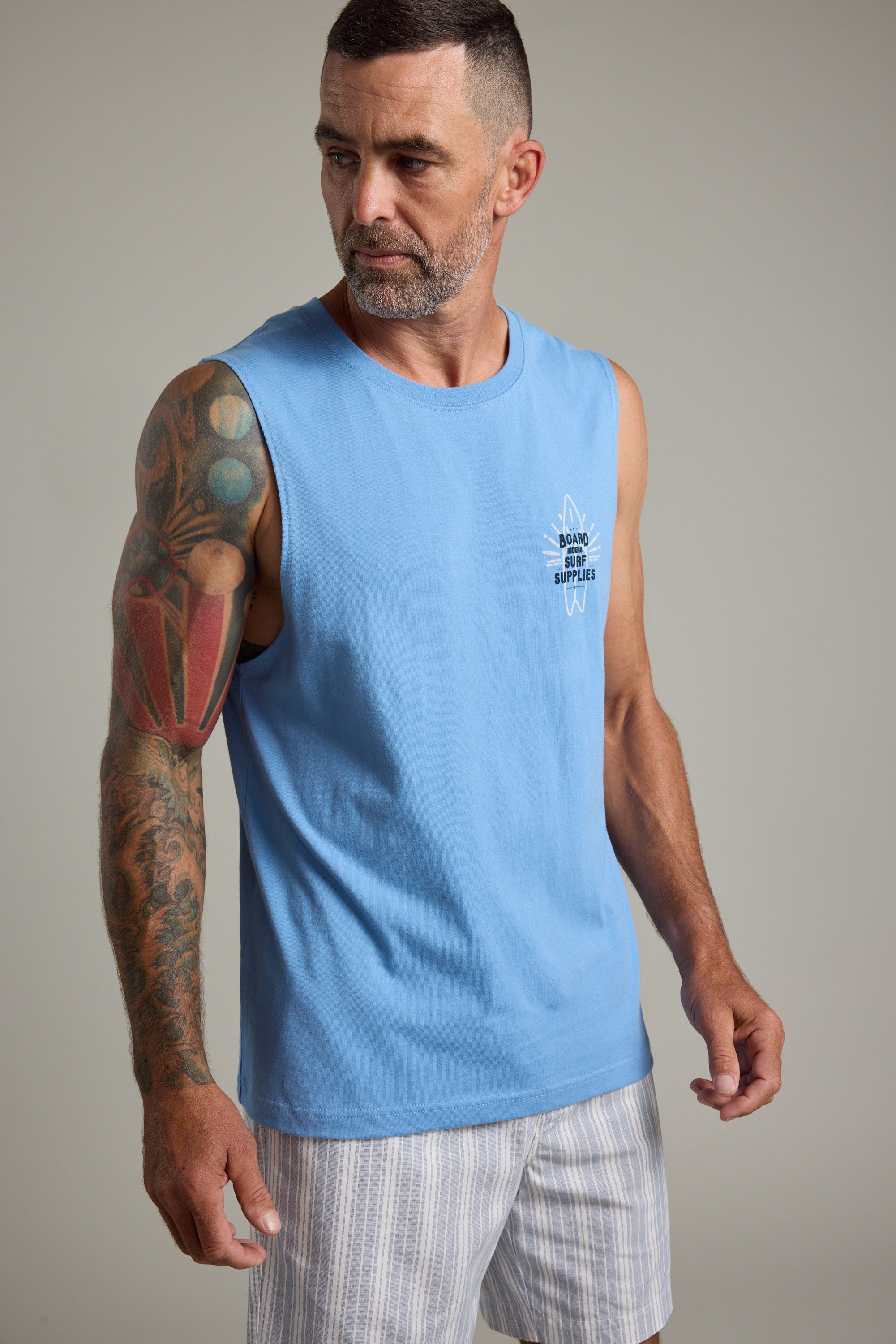 A man with a beard and colorful arm tattoo wears the Barkers BMC Board Riders Print Singlet and striped shorts, standing against a plain background and gazing slightly to the side.
