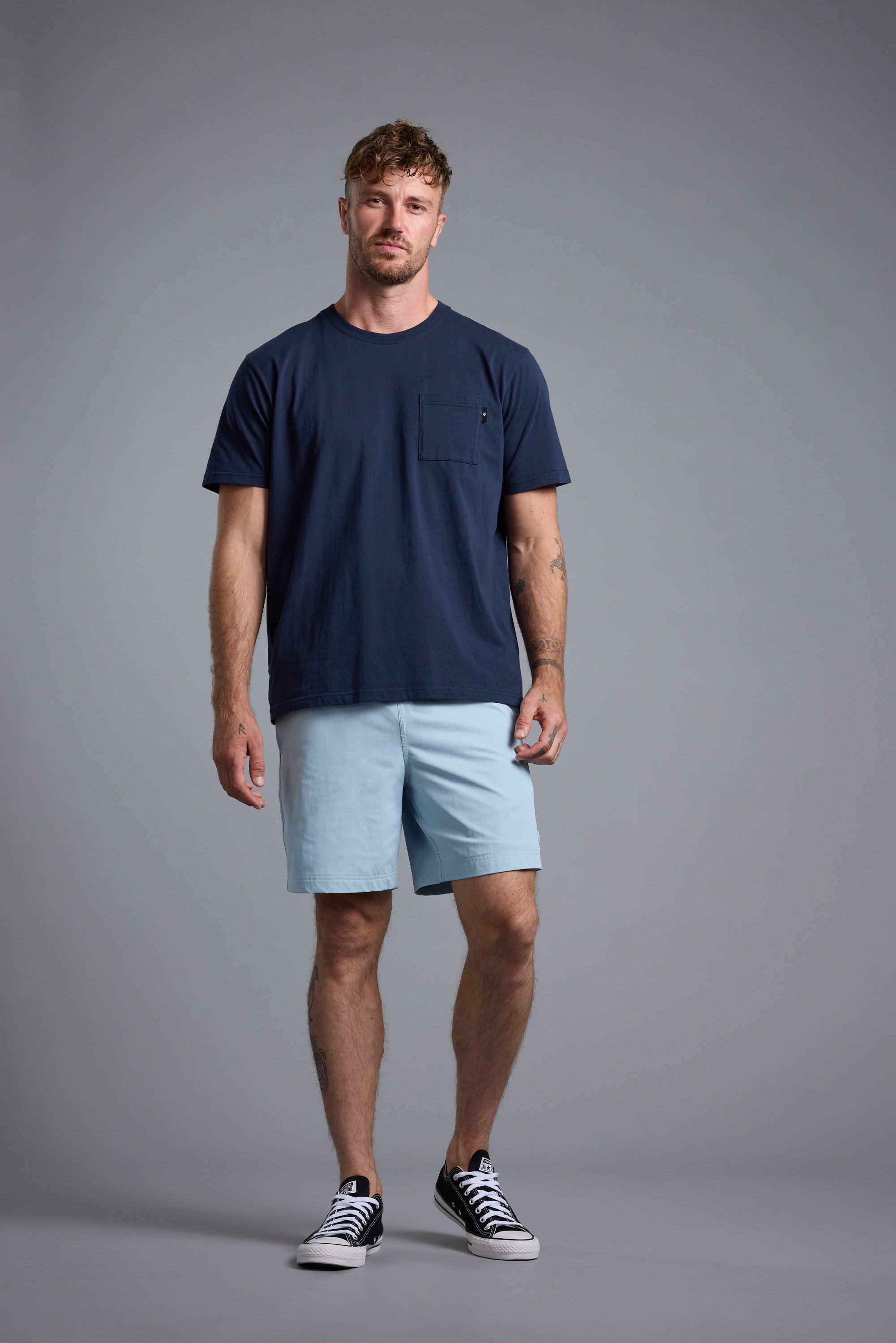 A man with short, curly hair and a relaxed expression stands against a plain gray background, wearing Barkers’ Summer Soul Sessions Print Tee in navy blue, paired with light blue shorts and black-and-white sneakers.