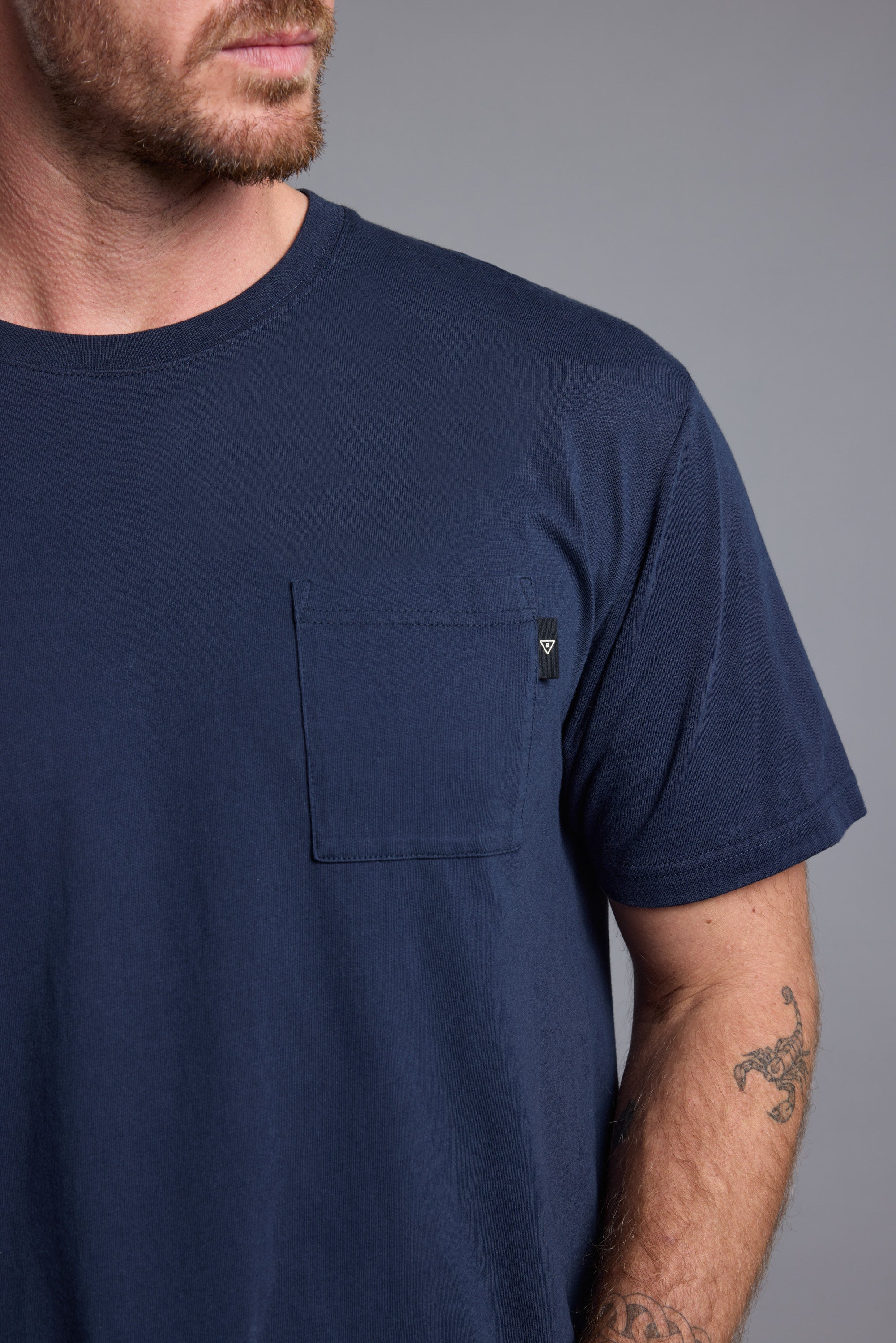 A man with light facial hair wears a Barkers Summer Soul Sessions Print Tee in navy blue, featuring a chest pocket. He shows part of his face and tattooed forearm against a plain gray background.