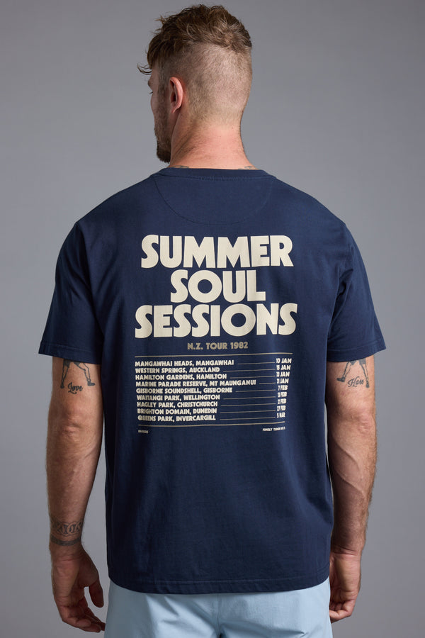 A man wears a Barkers Summer Soul Sessions Print Tee in navy blue organic cotton, featuring "SUMMER SOUL SESSIONS N.Z. TOUR 1982" and bold tour dates on the back, standing against a neutral gray background.