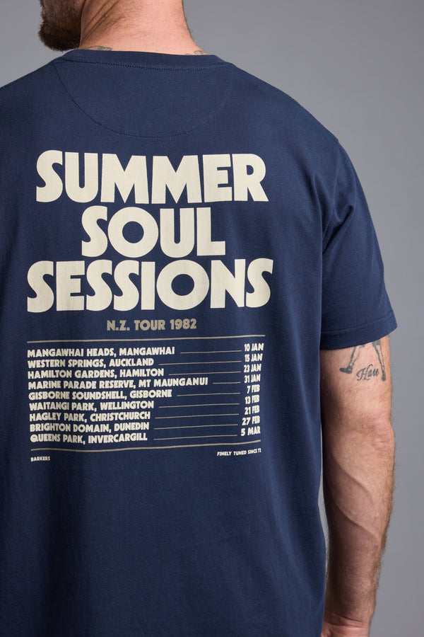 A person wears a Barkers Summer Soul Sessions Print Tee in navy blue organic cotton, with "SUMMER SOUL SESSIONS N.Z. TOUR 1982" and New Zealand concert dates in white on the back. A tattoo is visible on the person's arm.