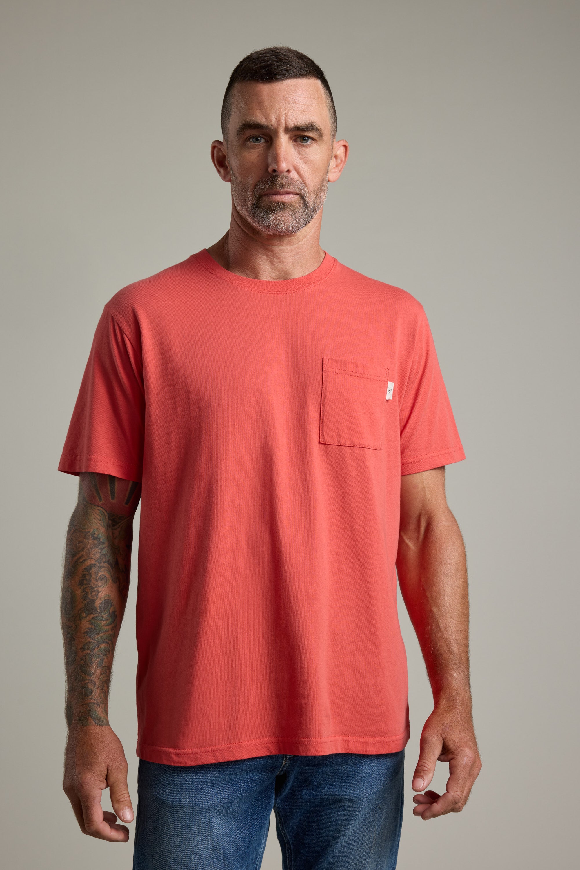 A man with short dark hair and a trimmed beard wears the Barkers Summer Soul Sessions Print Tee and blue jeans. He stands facing forward against a plain light background, displaying a tattoo on his left arm.