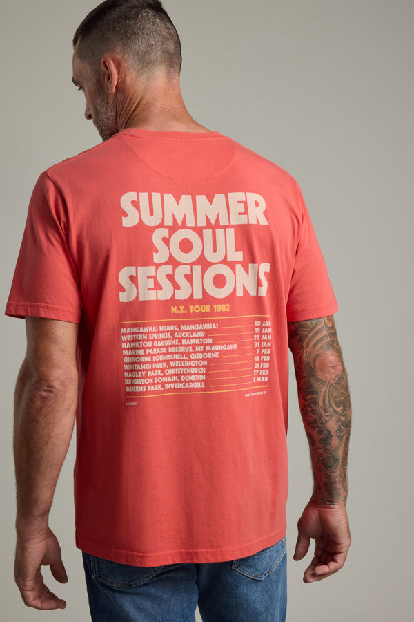 A man with short hair and a tattooed right arm wears a coral Barkers Summer Soul Sessions Print Tee, made from organic cotton, featuring “Summer Soul Sessions N.Z. Tour 1982” and tour dates in white on the back. He faces away from the camera.
