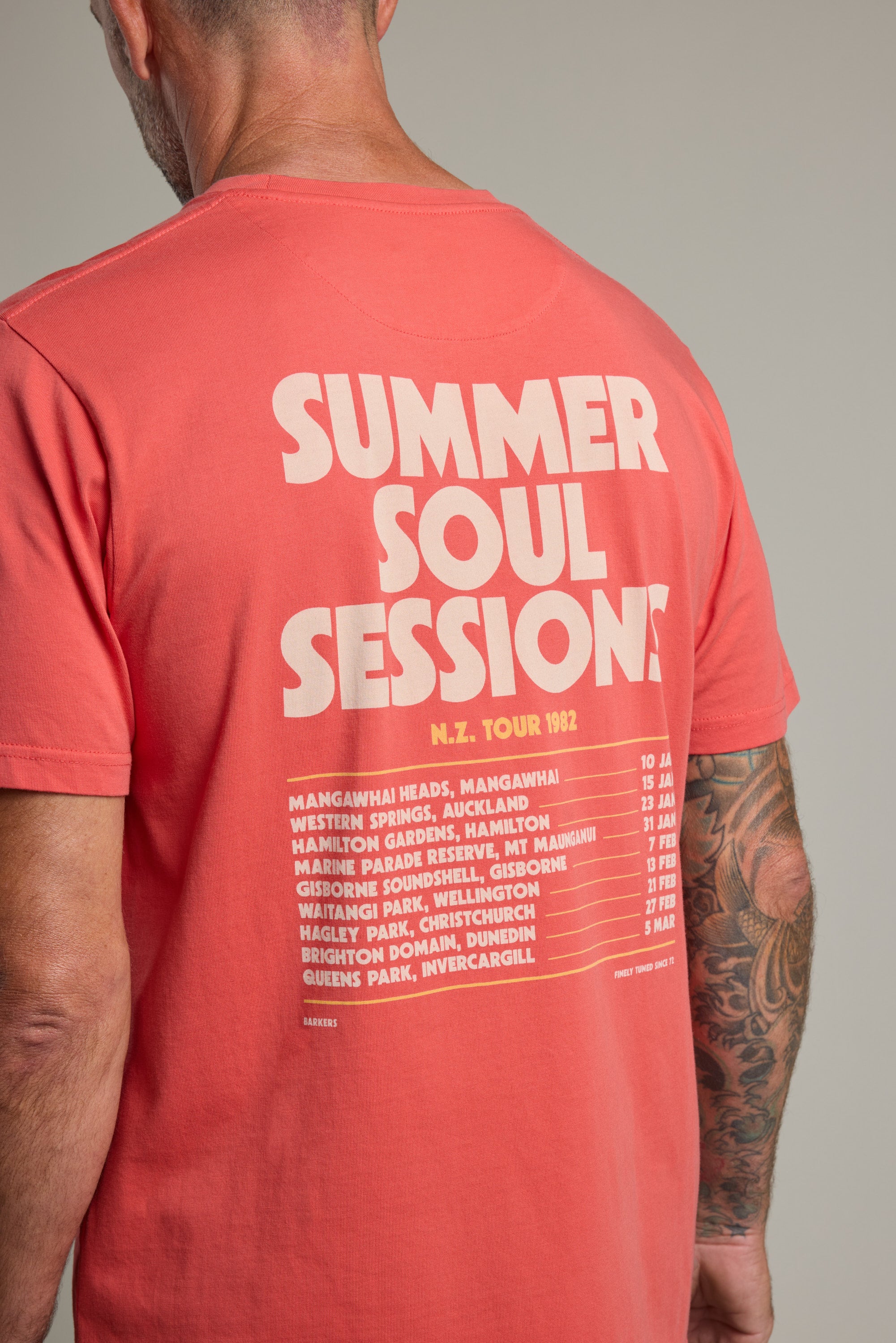 A man wears the Barkers Summer Soul Sessions Print Tee in coral, made from organic cotton, featuring "SUMMER SOUL SESSIONS N.Z. TOUR 1982" and New Zealand tour locations boldly printed in cream on the back.