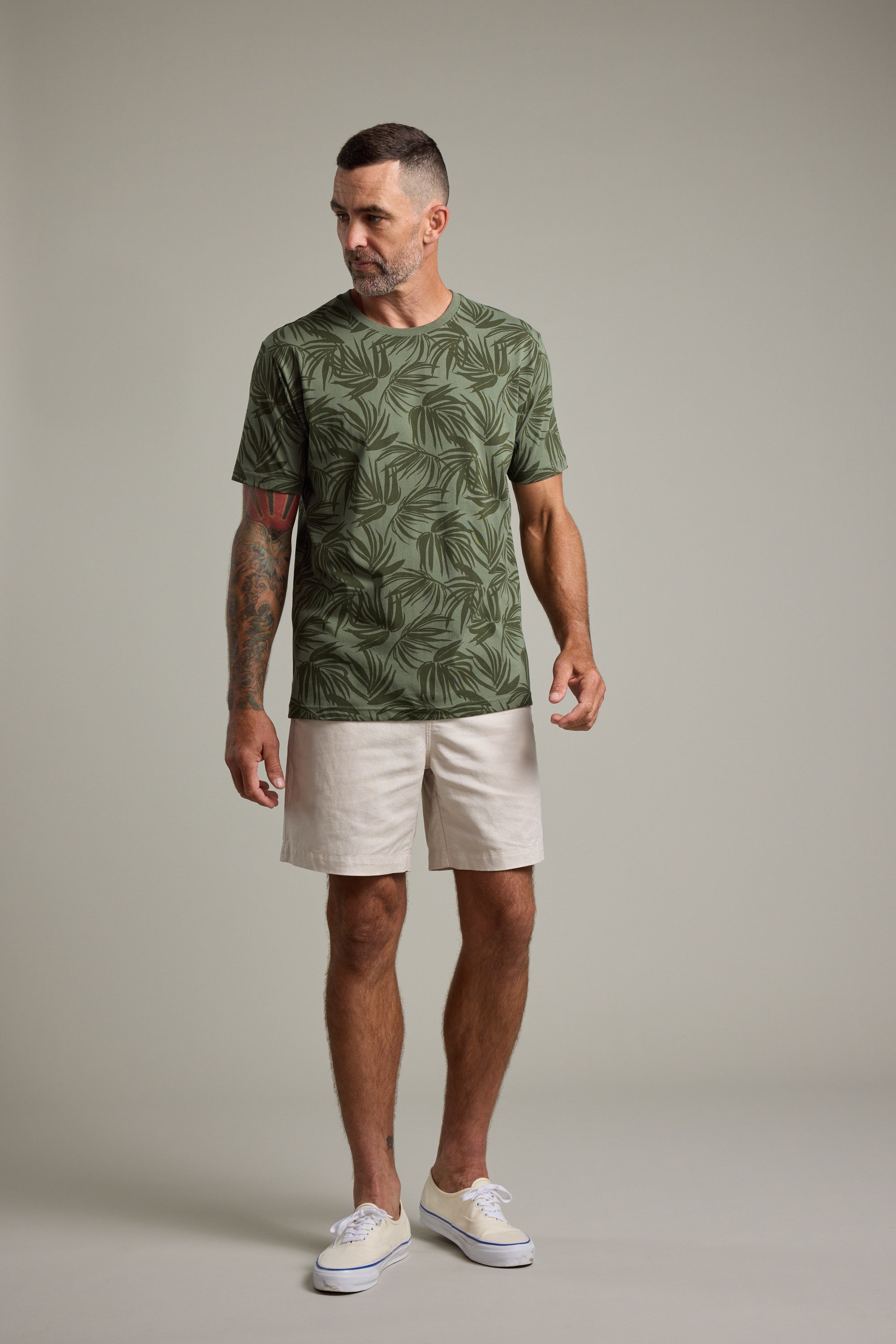 A man with short hair and a beard, tattoos on his left arm, stands against a plain background wearing the Barkers Palm Leaves Print Tee, light beige shorts, and white sneakers. He looks slightly to his right.