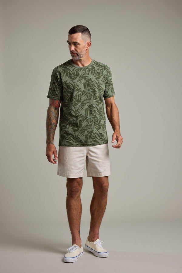 A man with short hair and a beard, tattoos on his left arm, stands against a plain background wearing the Barkers Palm Leaves Print Tee, light beige shorts, and white sneakers. He looks slightly to his right.