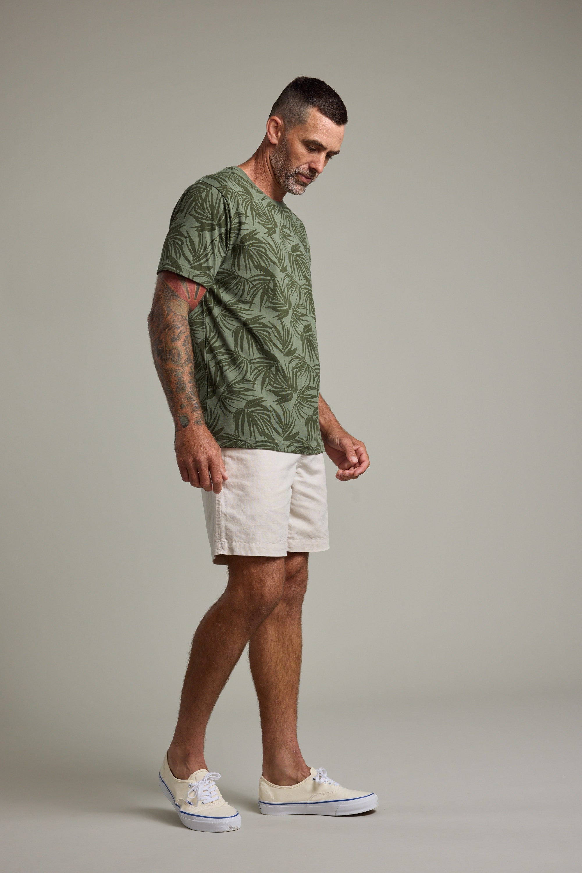 A man with short dark hair and tattoos on his right arm wears a Barkers Palm Leaves Print Tee, light shorts, and white shoes, standing against a plain light-gray background.