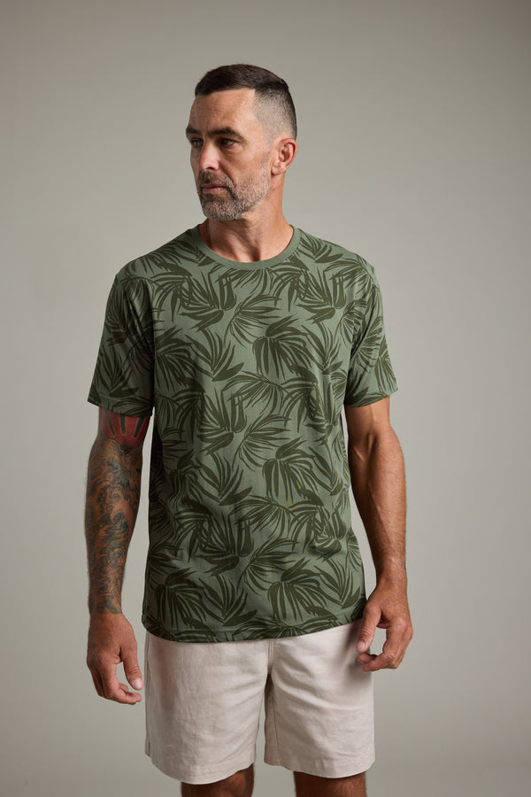 A man with short dark hair and a trimmed beard wears the Barkers Palm Leaves Print Tee and light shorts, standing against a plain gray background. He has a tattoo sleeve on one arm and is looking slightly to his left.