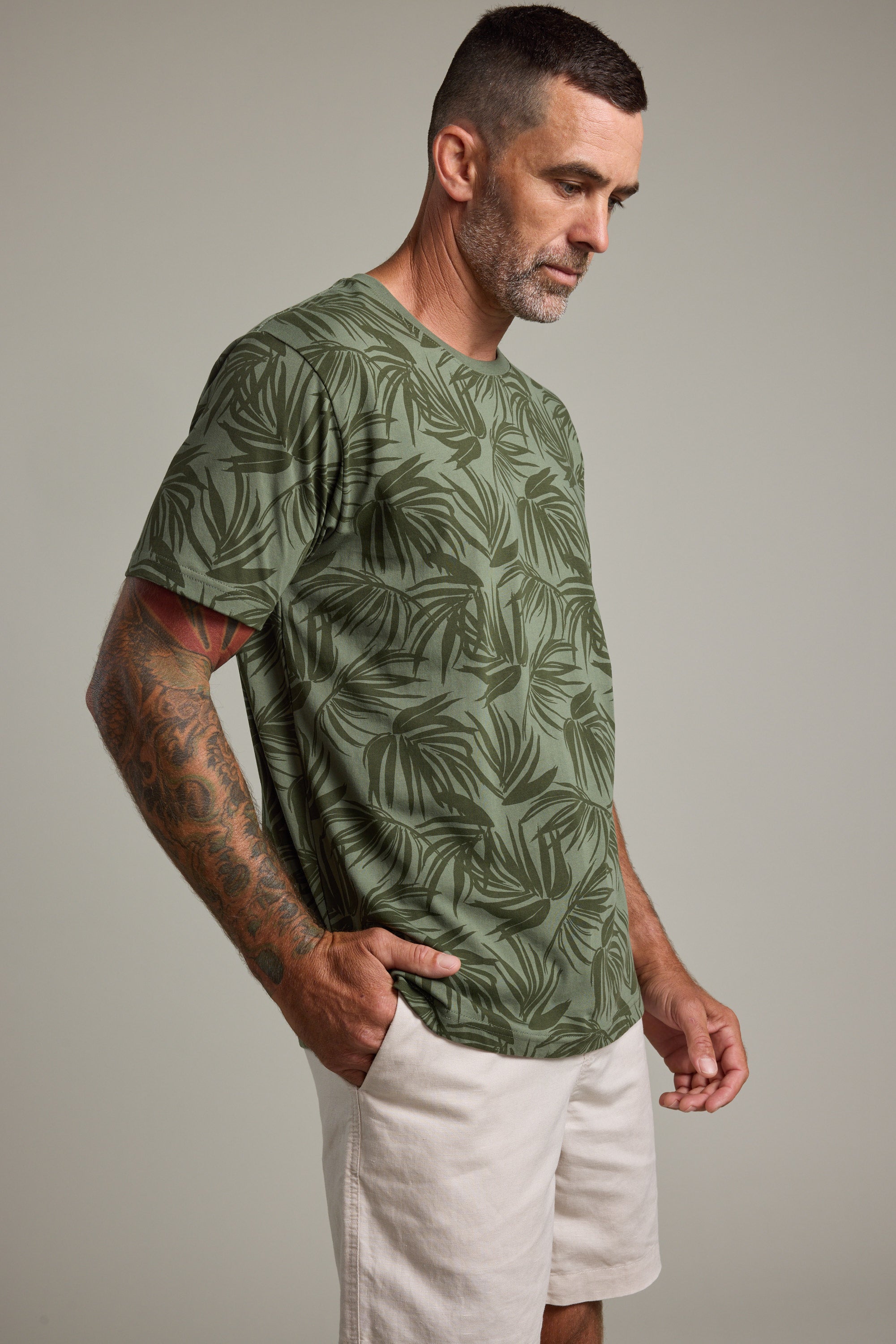 A man with short dark hair and a beard, wearing a Barkers Palm Leaves Print Tee and light shorts, stands with his hands in his pockets looking down against a neutral background.