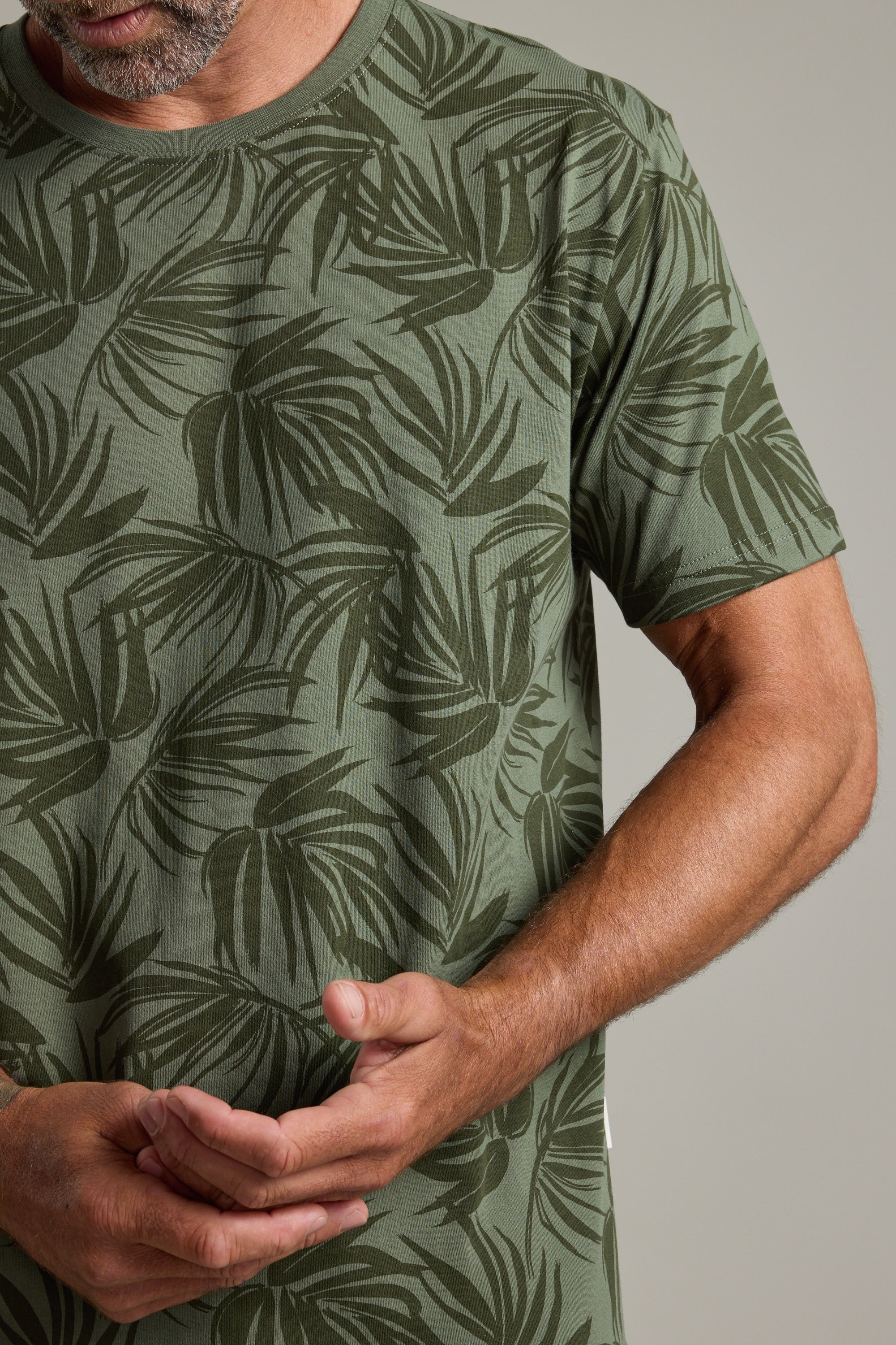 A man in a green Barkers Palm Leaves Print Tee stands against a plain background, his left arm bent and right hand touching his left palm.