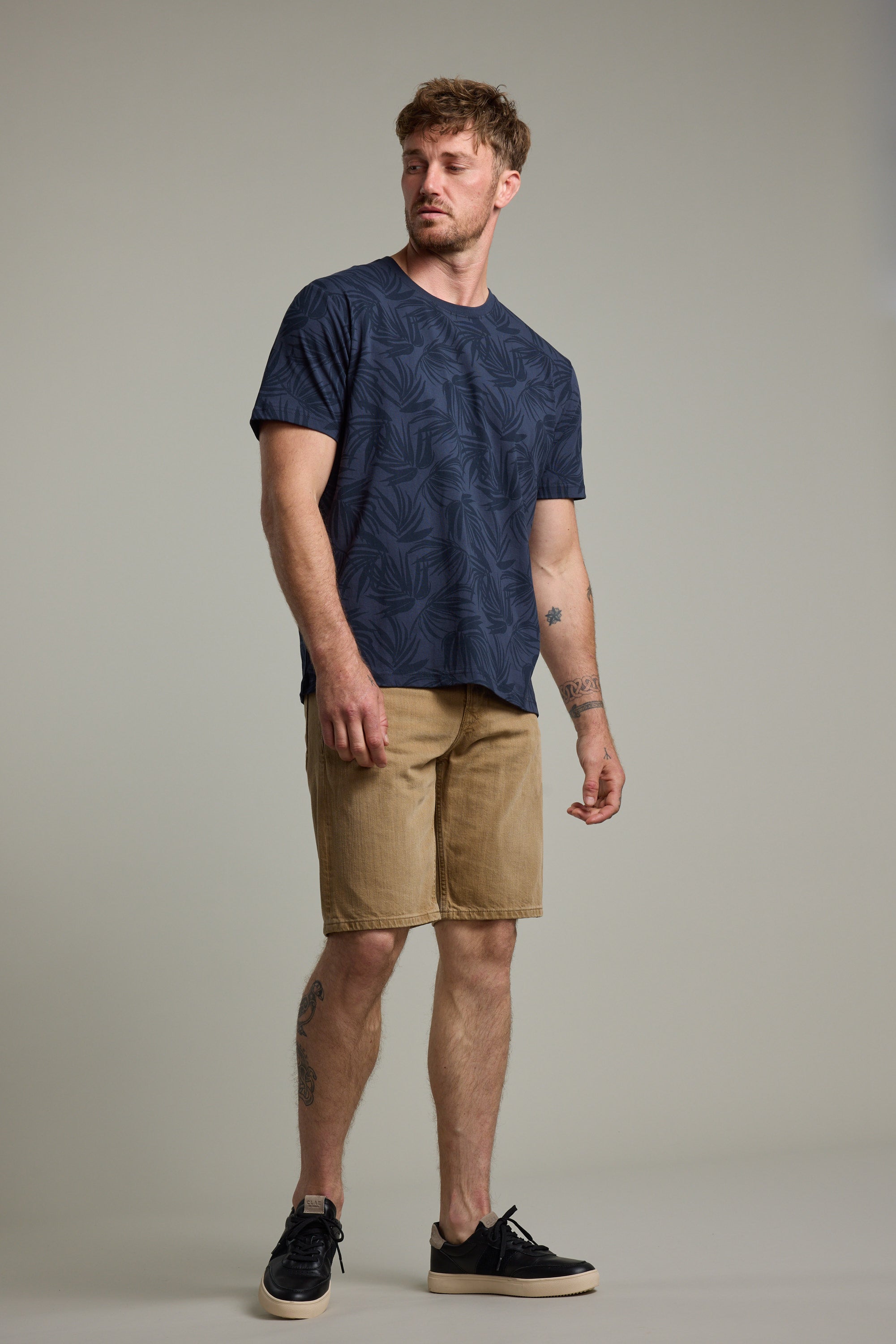 A man with short hair and tattoos poses against a plain background, wearing the Barkers Palm Leaves Print Tee in navy blue, tan shorts, and black sneakers. He stands relaxed, slightly looking to his left.