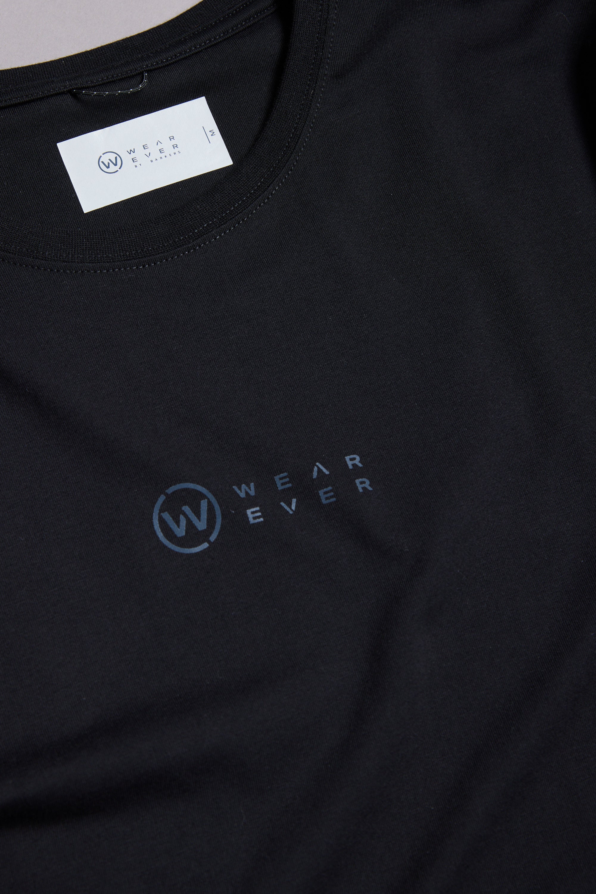 A close-up of the WearEver Outbound T-Shirt, a black cotton stretch tee featuring the dark blue WearEver logo on the chest and a white branded label inside the neckline.