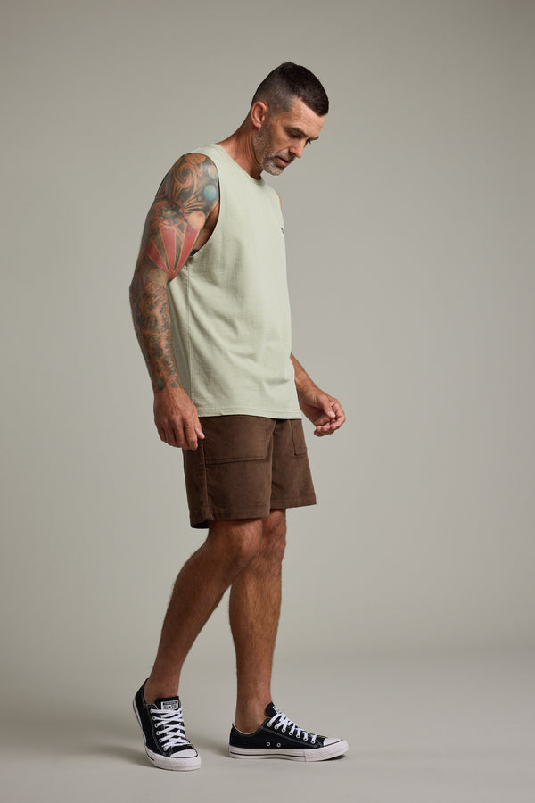 A man with short hair and tattooed arms wears the Barkers Newton Washed Singlet, styled with brown shorts and black sneakers. He stands against a plain background, looking down with a neutral expression.