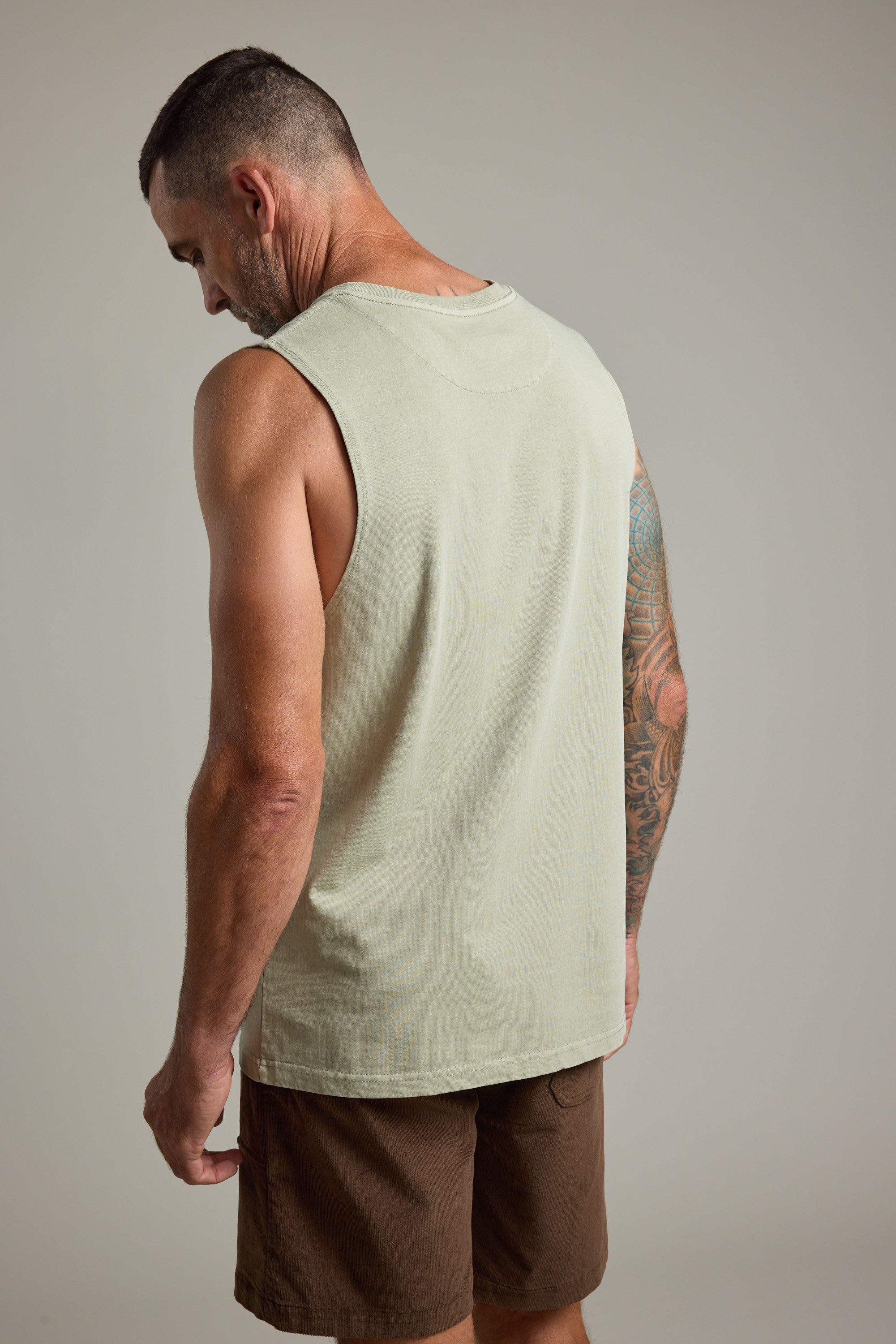 A man with short hair wears the Barkers Newton Washed Singlet in light beige and brown shorts, facing away to reveal his tattooed right arm against a plain background.