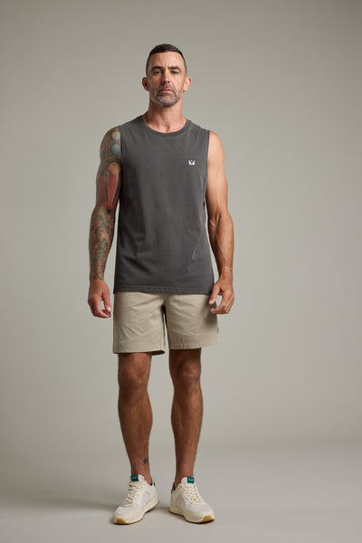 A man with short dark hair and a trimmed beard faces forward in a Barkers Newton Washed Singlet, dark gray, paired with beige shorts and white sneakers. He has colorful right-arm tattoos, a neutral expression, and stands against a plain background.