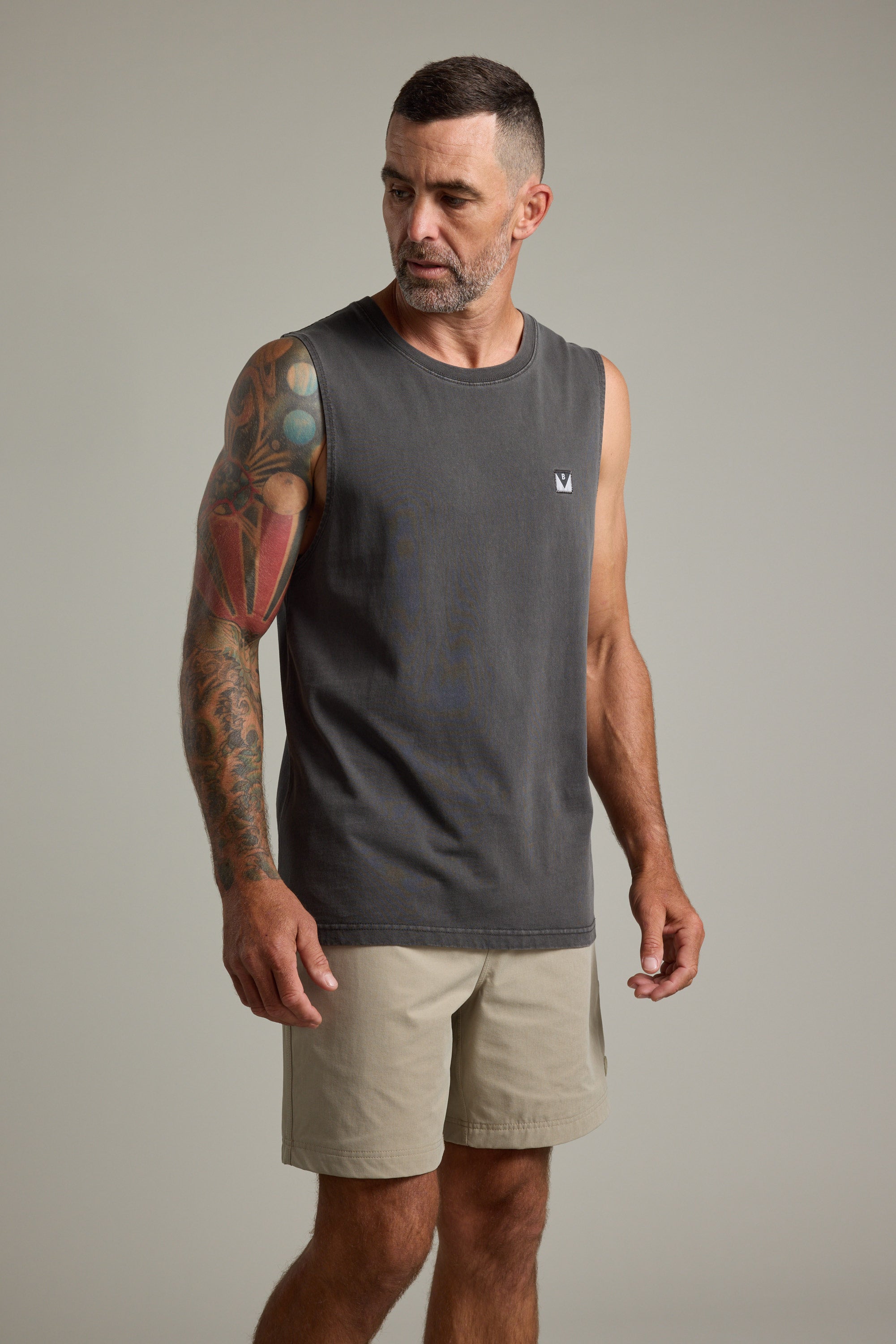 A man with short hair and a beard wears the Barkers Newton Washed Singlet in dark gray and beige shorts, revealing a colorful tattoo sleeve on his left arm. He stands against a plain backdrop, giving off a vintage vibe.