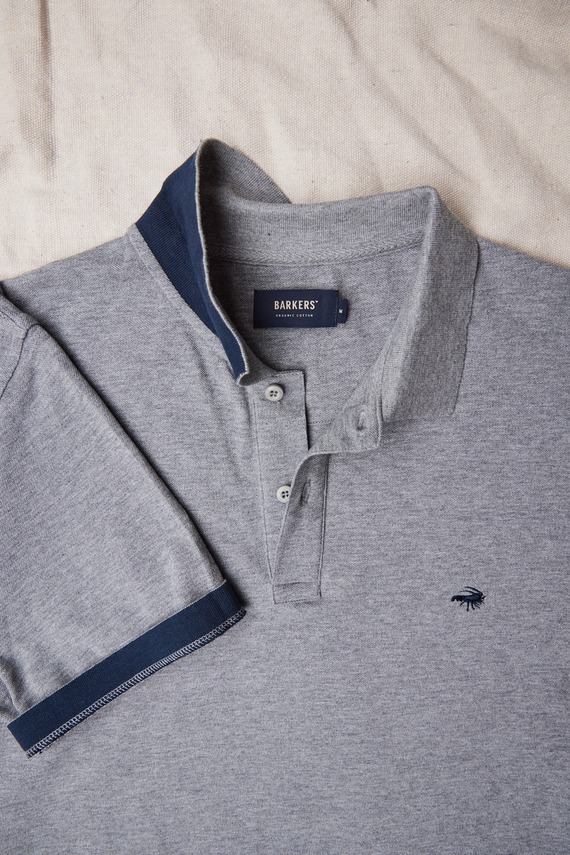 The Barkers Regent Pique Polo is a gray men's polo crafted from soft organic cotton, featuring a navy collar and sleeve trim, buttoned placket, and a small chest logo. It’s displayed flat on a light fabric surface.