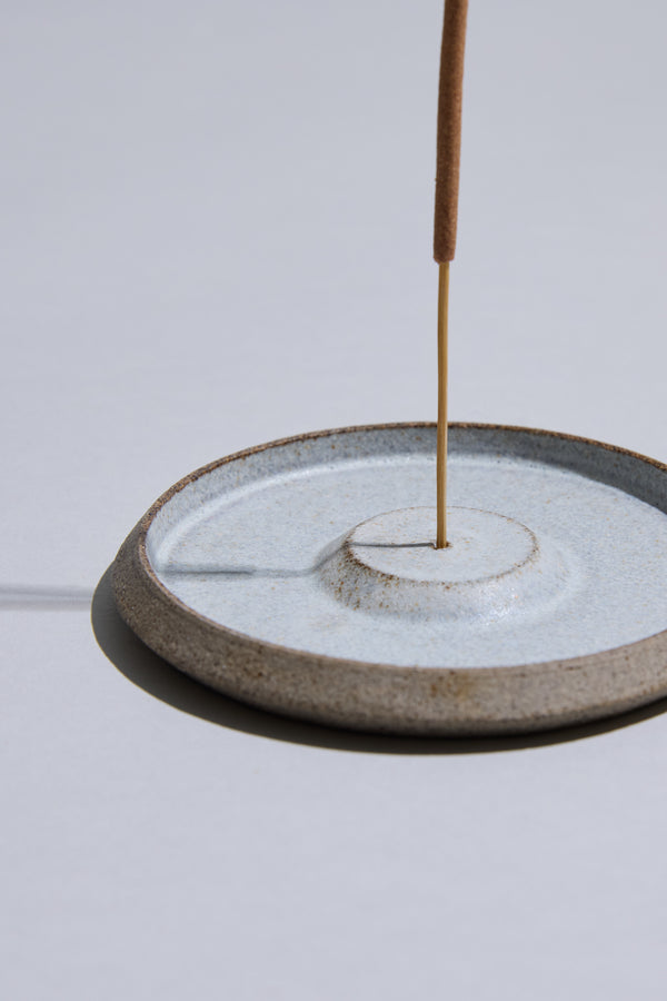 A brown incense stick stands upright in a round, light blue Studio Foon Incense Dish by Other, casting a shadow on a smooth, light-gray surface.