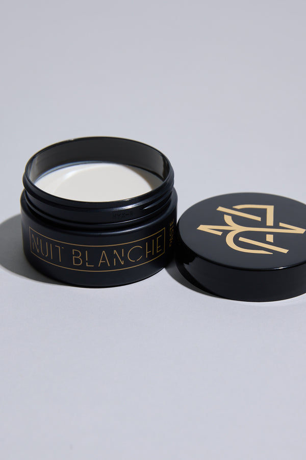 A black jar of Other's Nuit Blanche Paste with gold lettering, filled with white styling paste, sits on a light surface, its matching black and gold lid beside it.