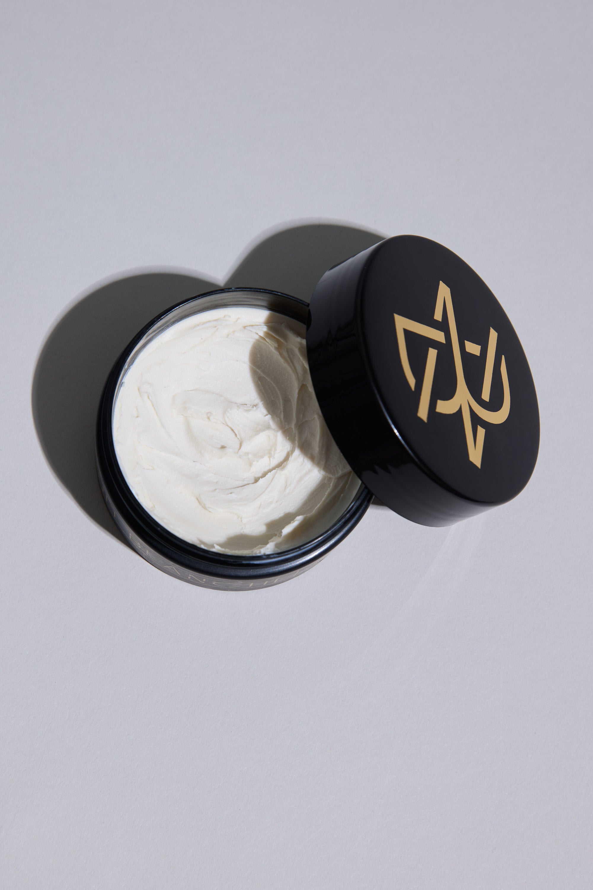 A black container of Other's Nuit Blanche Paste, featuring a gold geometric lid design and partially open to reveal smooth white styling paste inside, is displayed on a light gray surface.