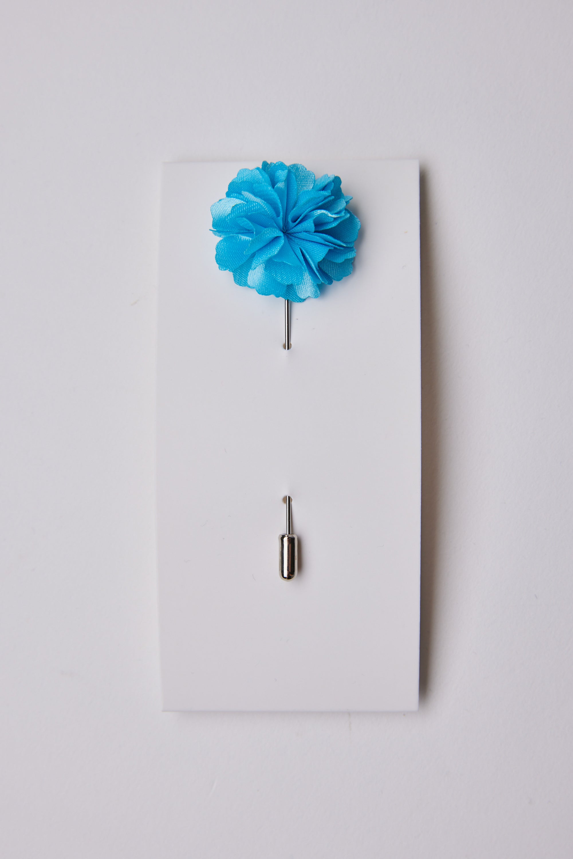 The Parisian Fraser Flower Lapel Pin, a stylish blue suiting accessory, is attached to a white card with its silver pin cap positioned below the flower.