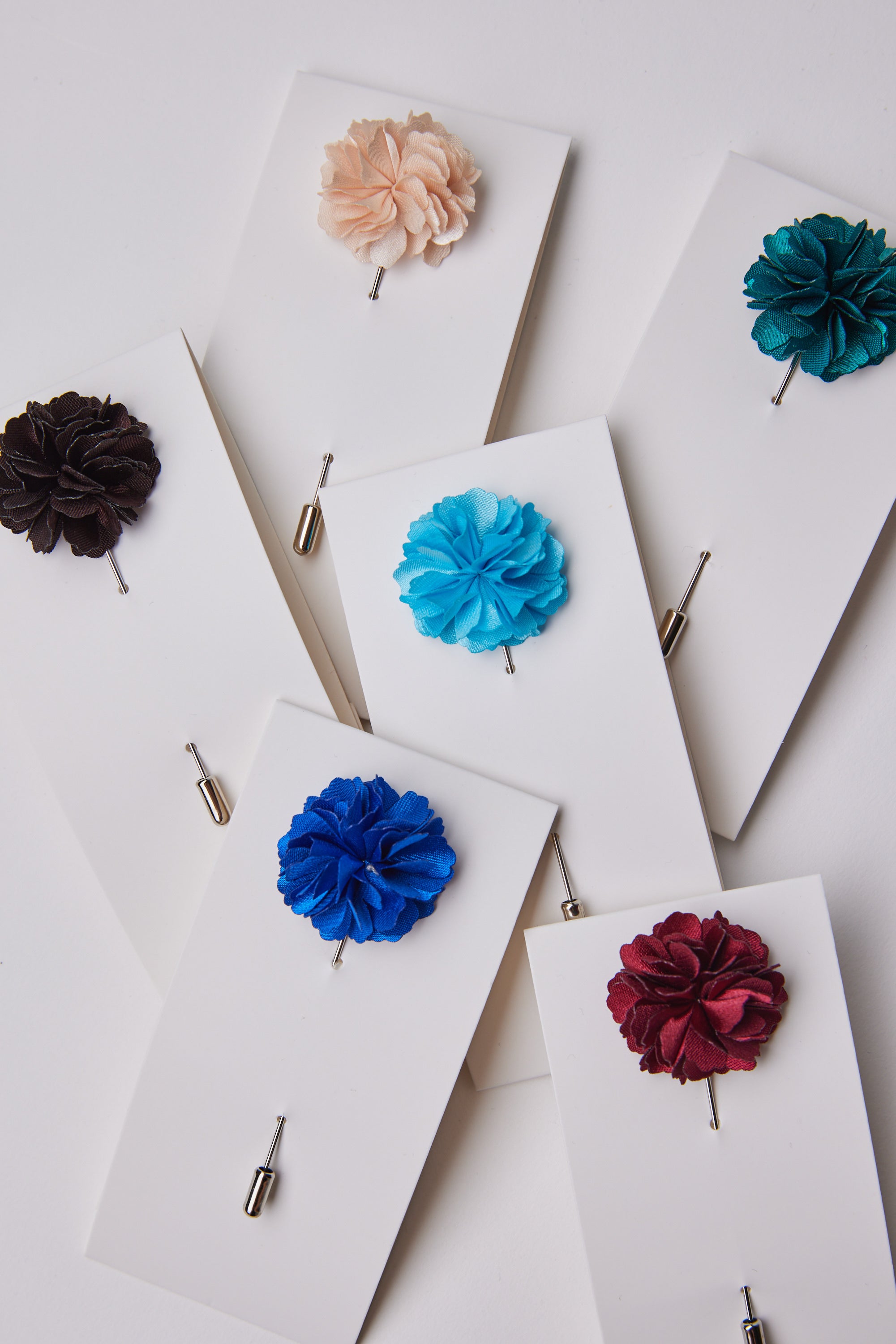 Parisian Fraser Flower Lapel Pins in black, beige, teal, turquoise, blue, red, and burgundy are displayed on white cards atop a white surface—an elegant accessory to enhance any formal outfit.