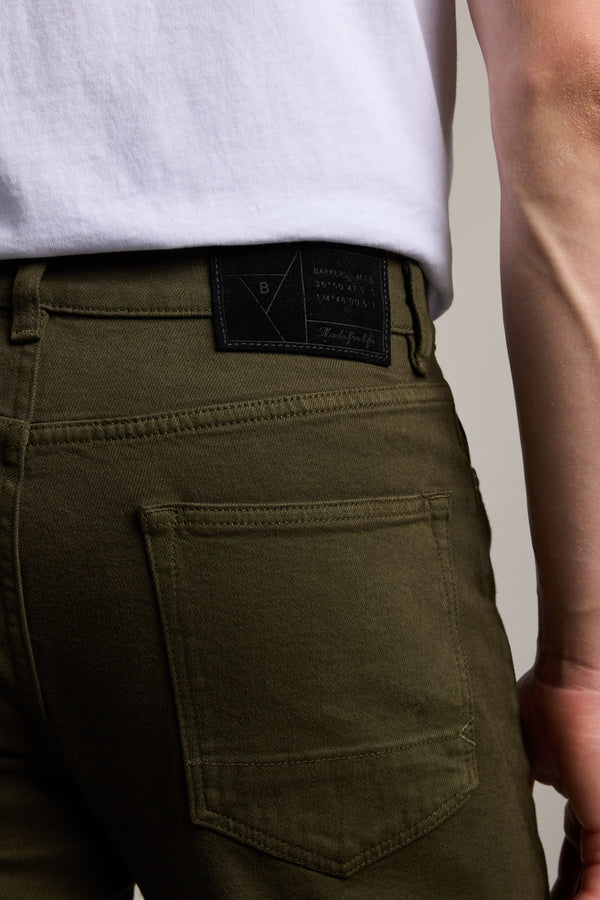 A person wearing Barkers Rambler Slim Coloured Jean in olive green, made with regenerative cotton and a white shirt, shown from the waist down with focus on the back pocket and a black Barkers label on the waistband.