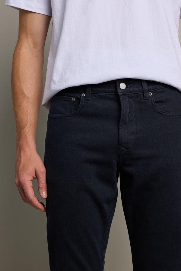 A person wears a white t-shirt and Barkers Rambler Slim Coloured Jeans in dark blue, shown from shoulders to mid-thigh with one arm relaxed by their side, set against a plain background. Crafted in Vietnam.