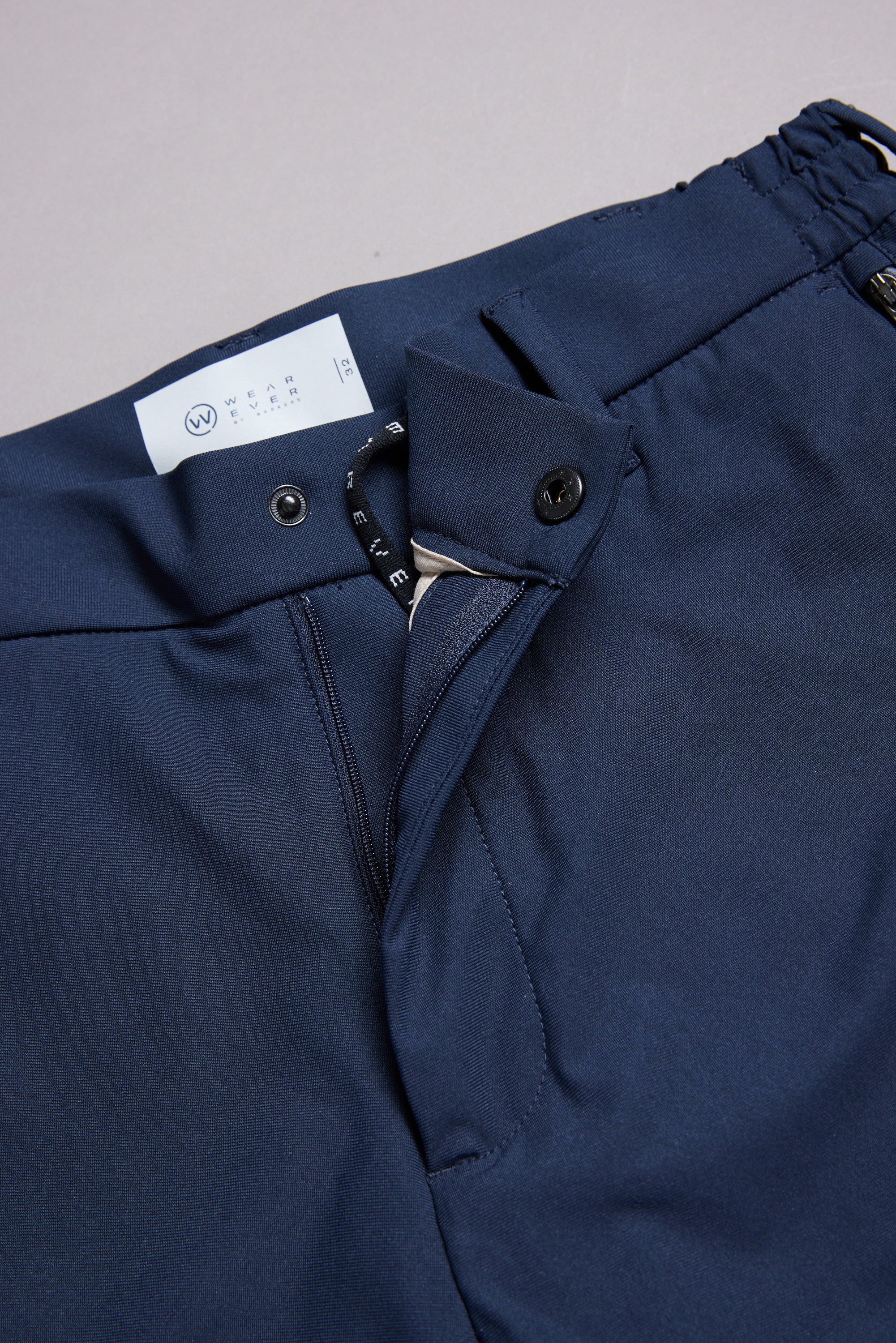 Close-up of WearEver Long Game Pant by WearEver: navy blue trousers with open waistband, displaying button, zipper, and label—highlighting chino-inspired style for comfortable, stylish travel.