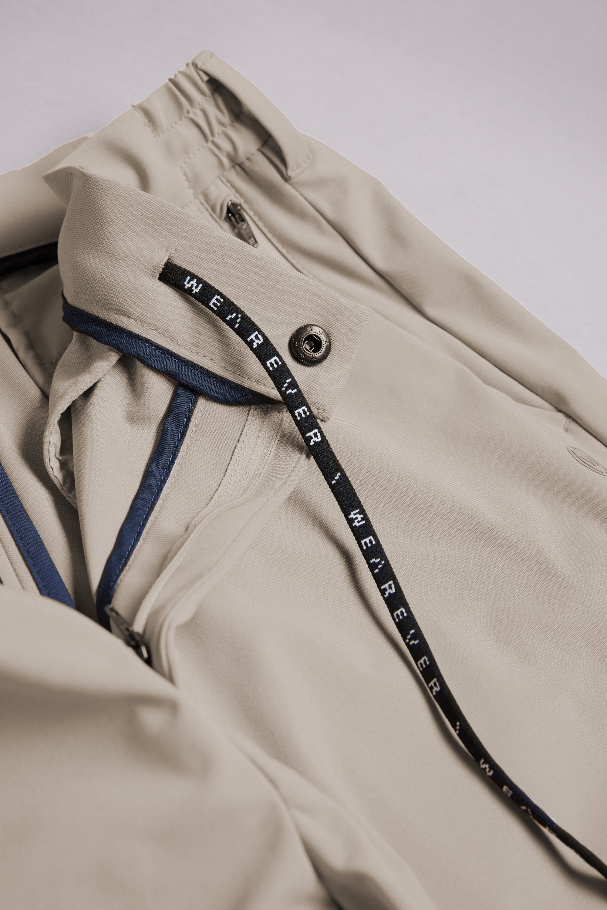 A close-up of the WearEver Switch Short by WearEver shows a beige jacket with a zipper, snap button, black “WE R E V E R” drawstring, navy blue inner seam trim, and comfort stretch for easy movement.
